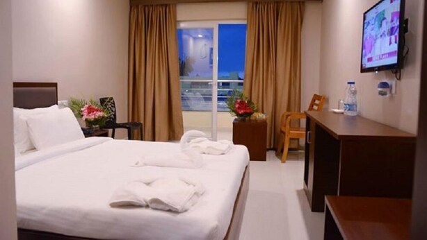 A hotel room with a bed, a desk, a chair, a TV, and a window with curtains. - Hotel Shree Hari Grand (Puri)