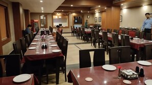 A dining area with multiple tables set for meals, a Coca-Cola refrigerator, and a person standing near the back. - Hotel Shree Hari Grand (Puri)