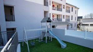 Un terrain de jeux avec un toboggan et des balançoires sur une pelouse artificielle, entouré d’un bâtiment blanc doté de balcons et de fenêtres.