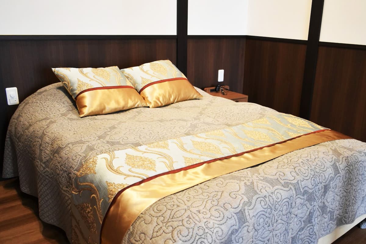 Ein ordentlich bezogenes Bett mit gemustertem Überwurf und zwei Kissen, ein Nachttisch aus Holz mit einer Lampe und ein dunkles Holz-Kopfteil.