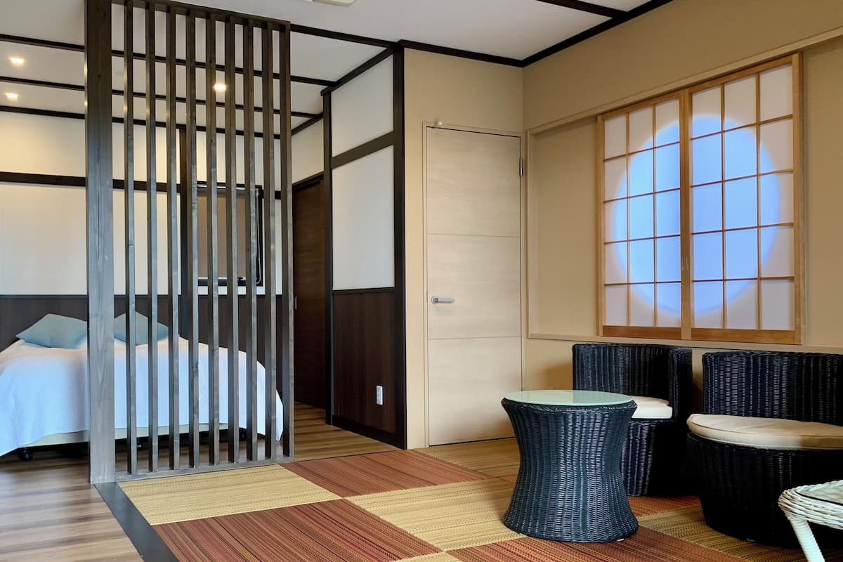 Ein Hotelzimmer mit Bett, einer Raumtrennung, einer traditionellen Schiebetür, einer Tatami-Matte und Blick auf eine Stadtlandschaft durch einen Shoji-Bildschirm.