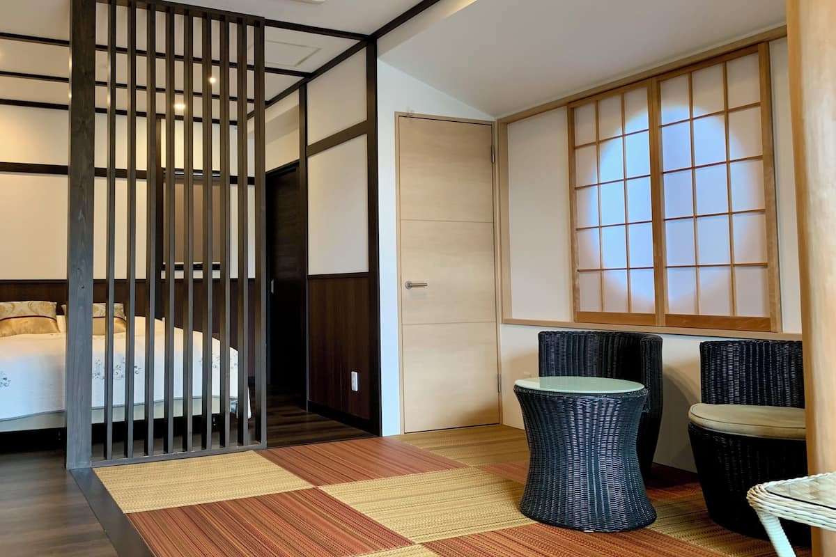 Habitación de hotel con cama, pared divisoria, estera de tatami, puerta corredera tradicional japonesa y una pequeña mesa con silla.
