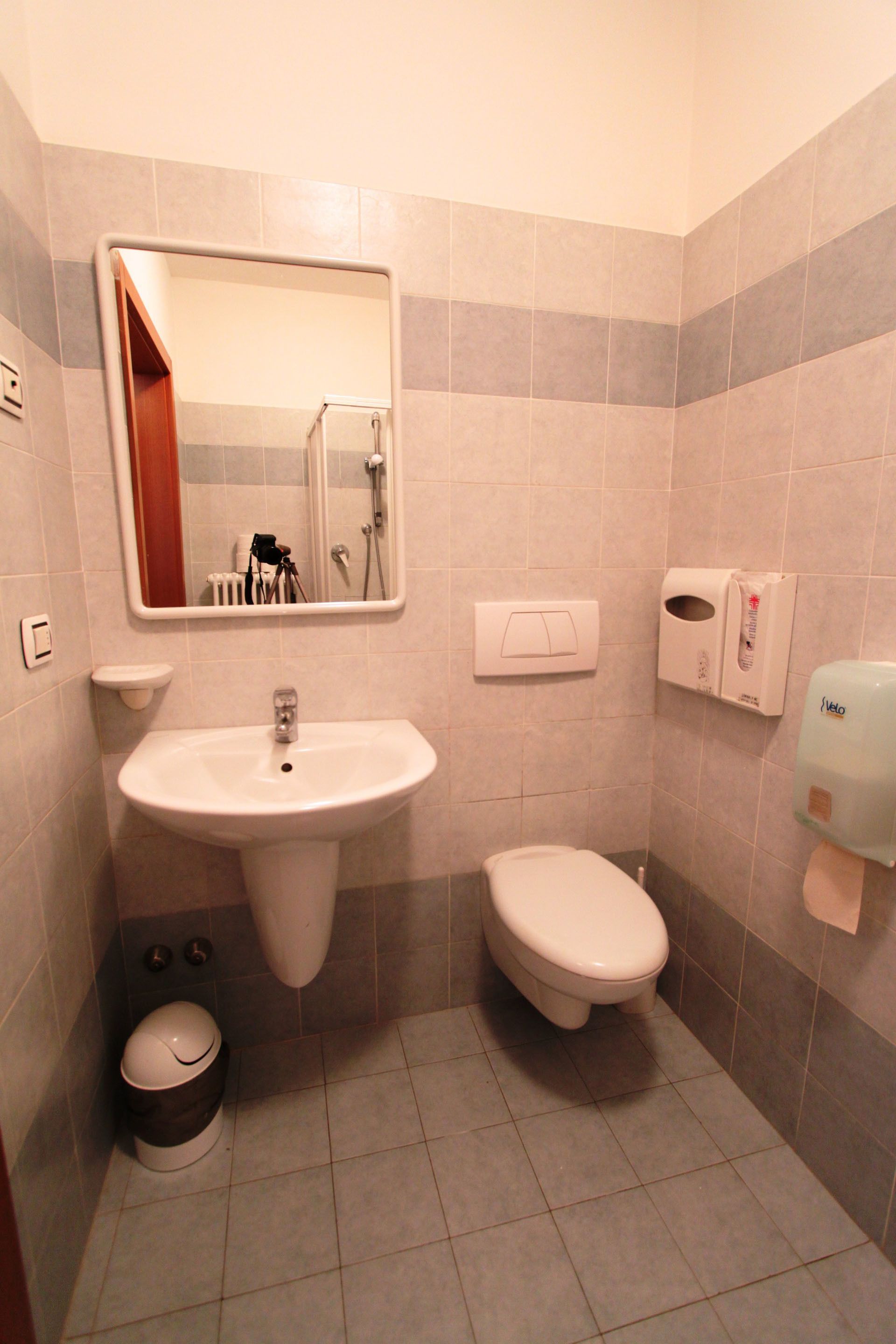A small bathroom with a sink, toilet, and a mirror. There is a soap dispenser and a trash can.