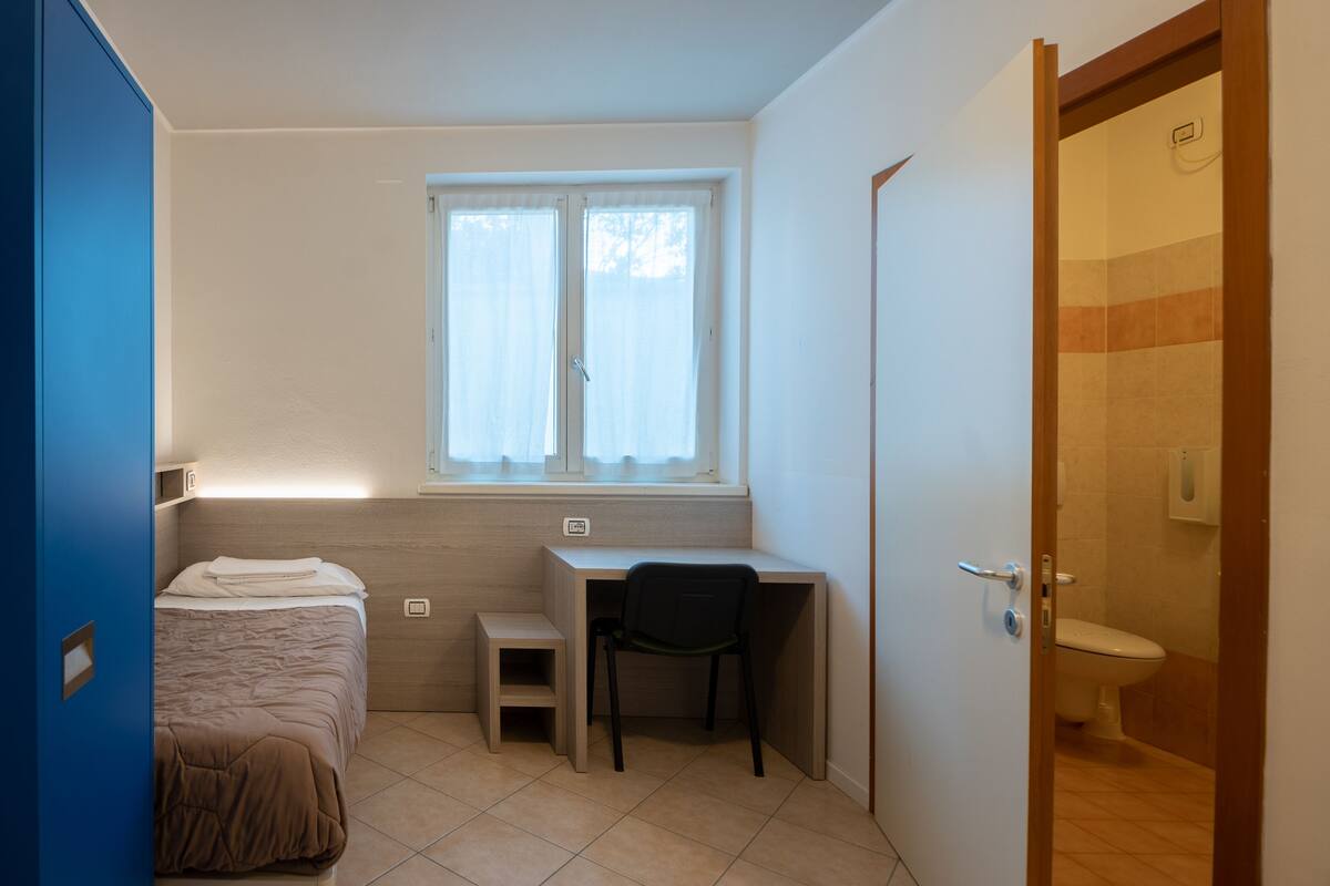 Ein kleiner, kompakter Raum mit Bett, Schreibtisch, Stuhl und Fenster.