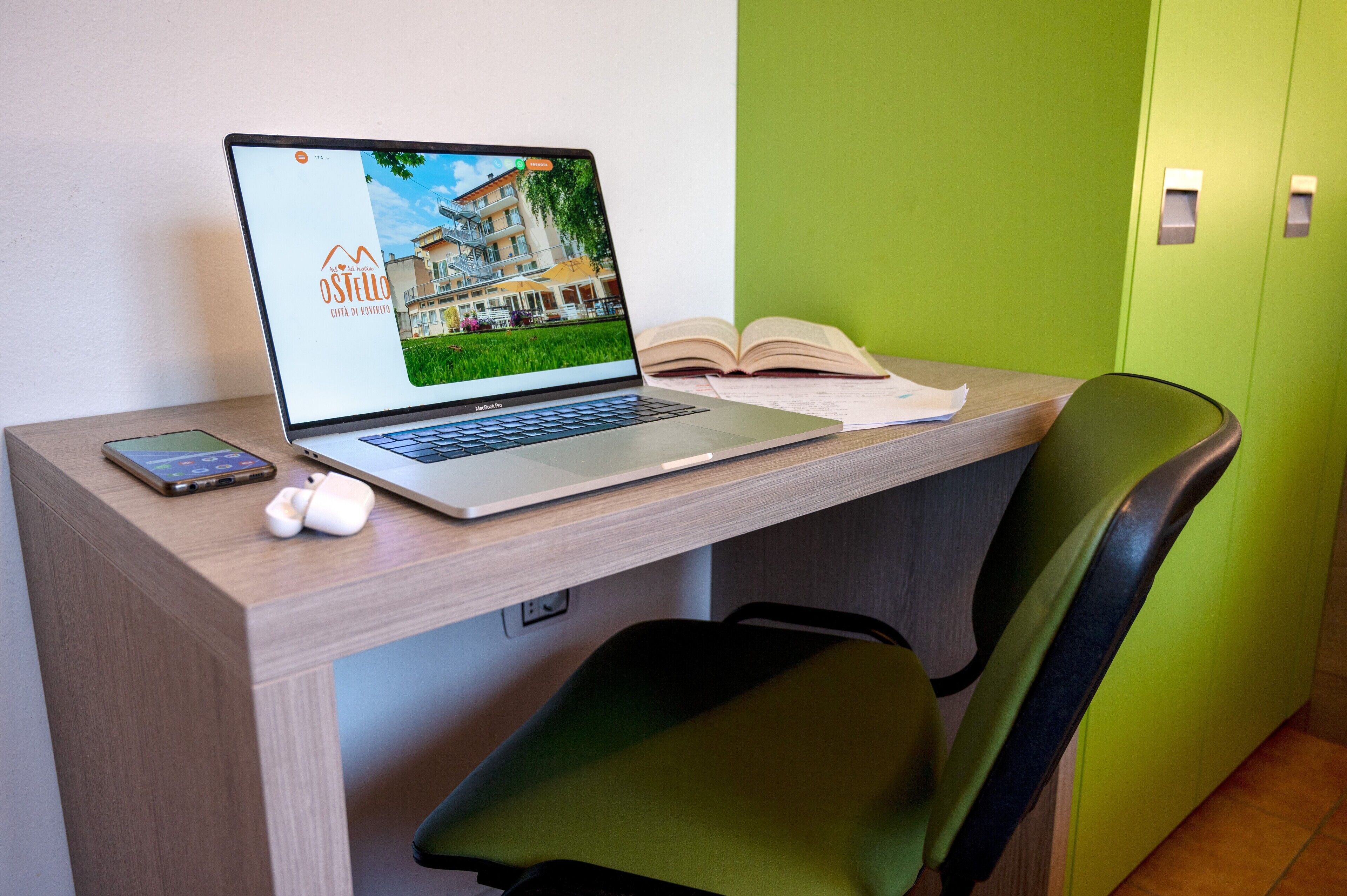 Una computadora portátil sobre un escritorio, junto con un teléfono inteligente, audífonos, un libro abierto y una silla verde.
