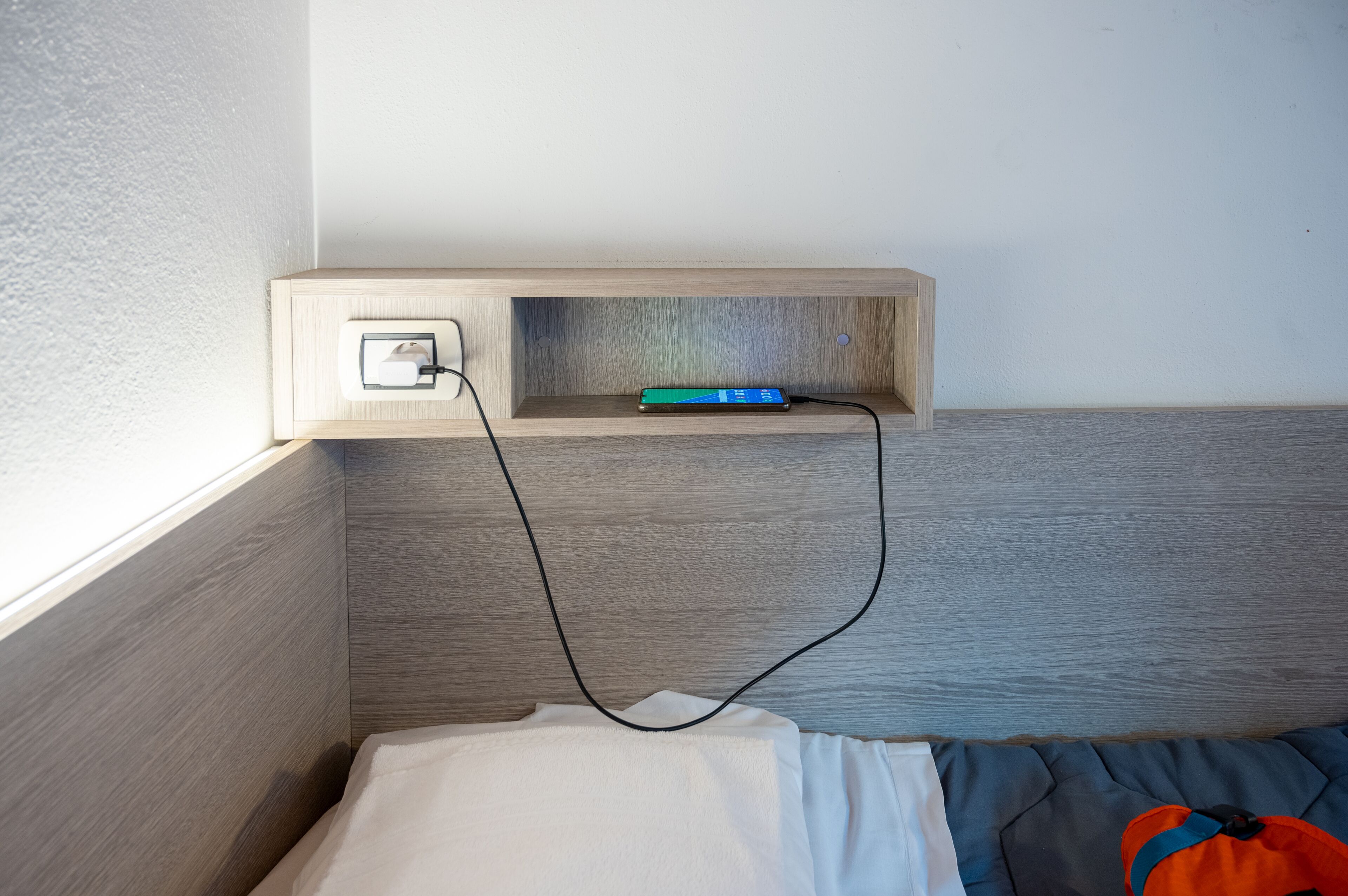 A bed with a white sheet, a wooden headboard, a small wooden shelf with a power outlet and a charging device, and a window with daylight coming through.
