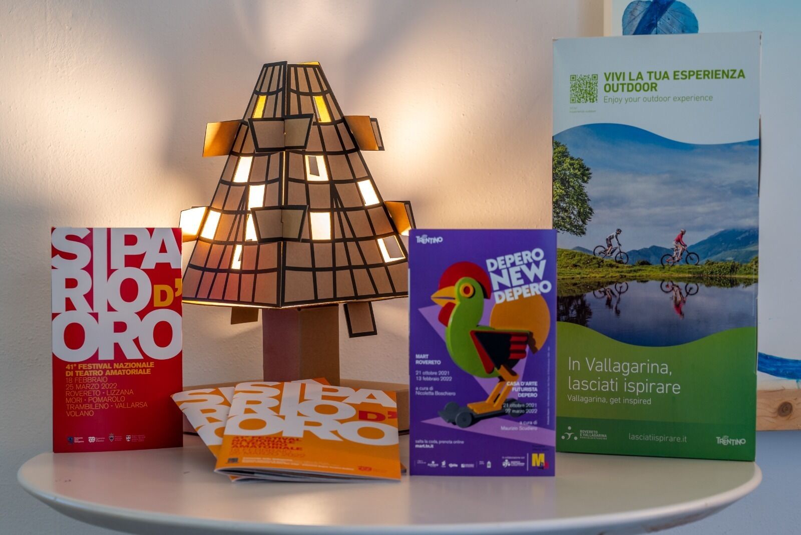 Una mesa con una lámpara, un libro rojo titulado 'SIPARI D'ORO', un libro morado titulado 'DEPERO NEW DEPERO' y un libro verde titulado 'VIVI LA TUA ESPERIENZA OUTDOOR'.