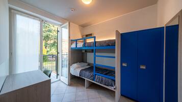 A dormitory room with bunk beds, a desk, and a sliding glass door to a balcony.
