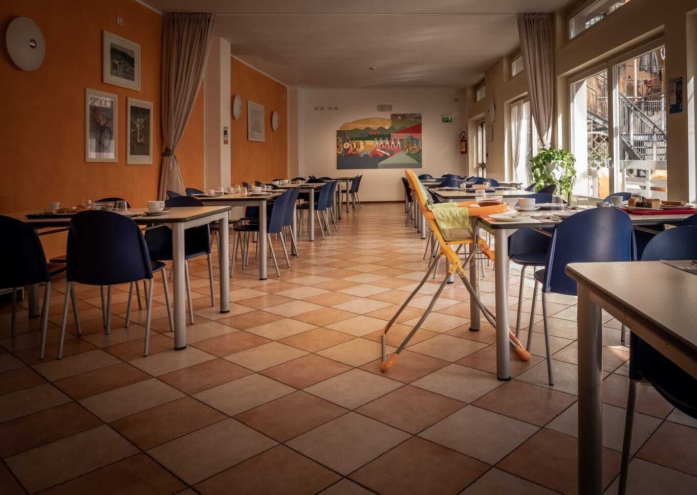 Uma sala de jantar com várias mesas e cadeiras, piso ladrilhado e um mural na parede.