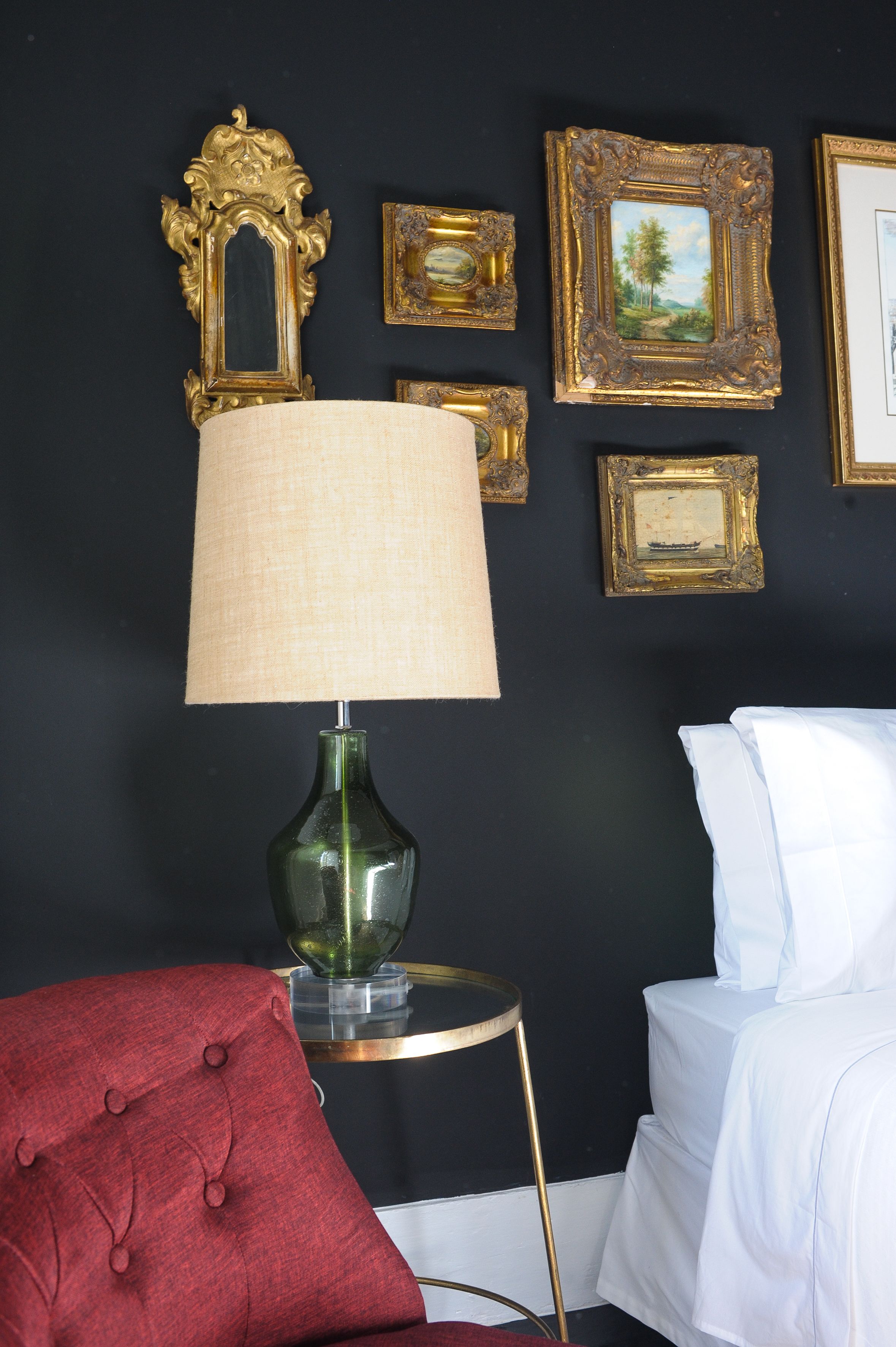 A room with a bed, a red upholstered chair, a green lamp, and several framed pictures on the wall.