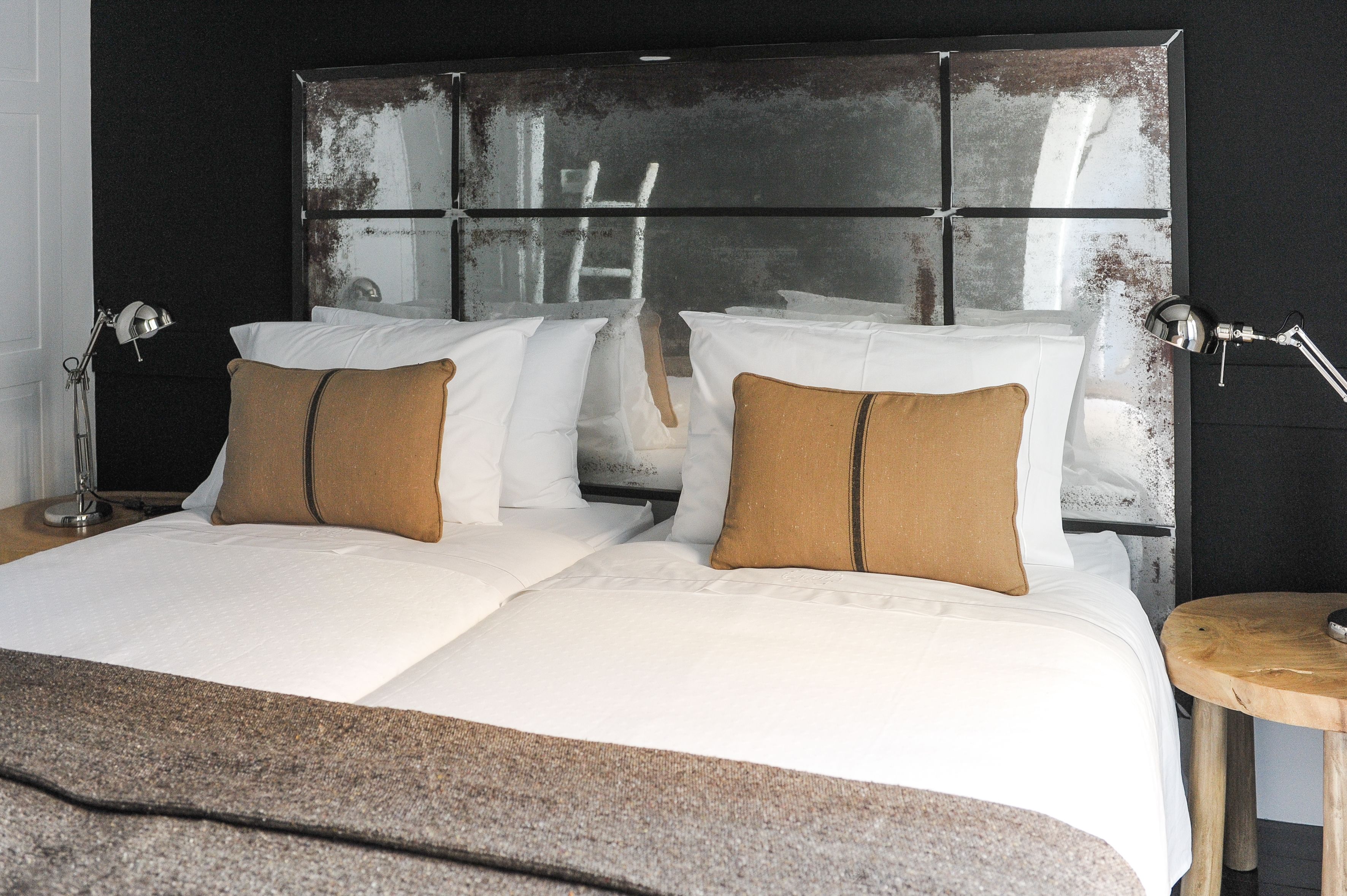 A double bed with white linens and two brown pillows, a bedside table with a lamp, and a large mirror with a distressed frame.