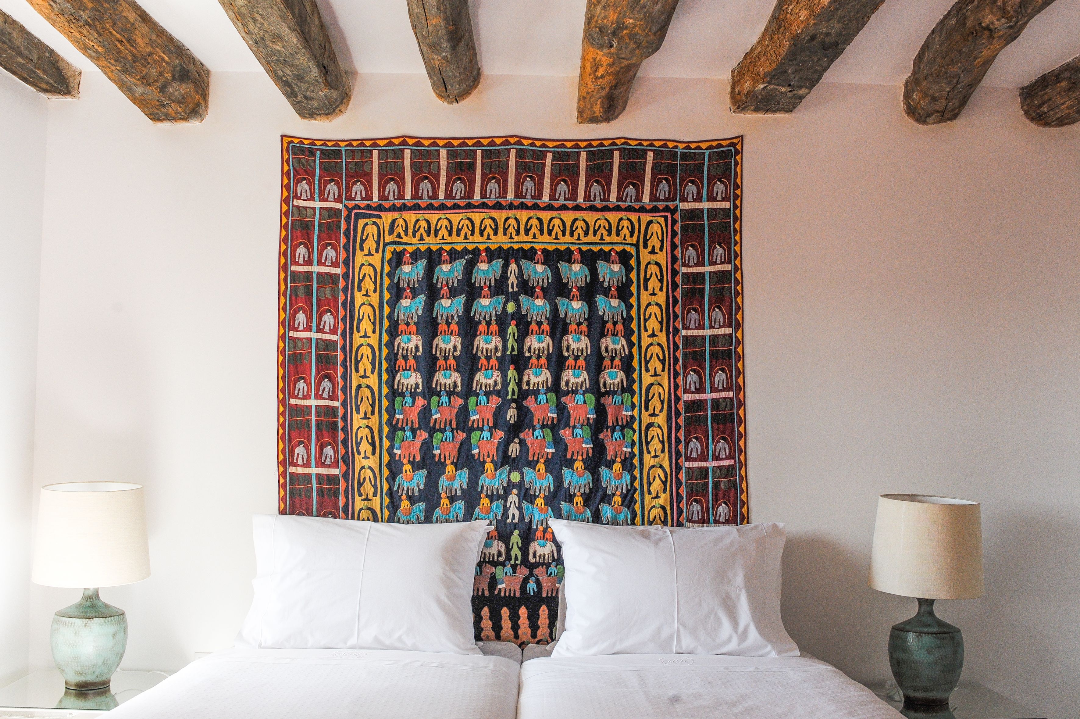 A bed with white linens, a tapestry with colorful patterns, and a wooden ceiling.