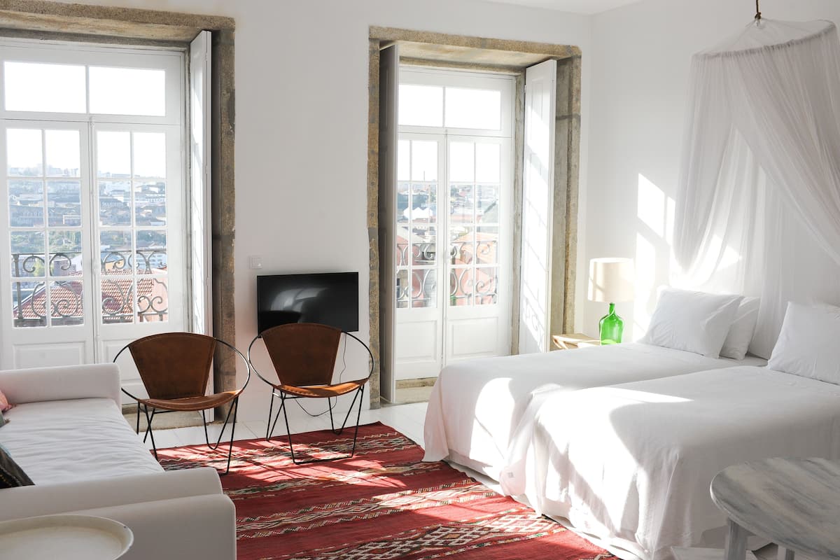 A hotel room with a bed, two chairs, a TV, and a view of the city through large windows.