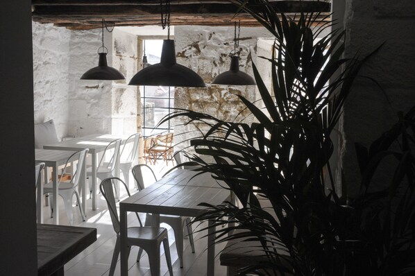 A rustic dining area with wooden tables and white chairs, large hanging pendant lights, and a potted plant.