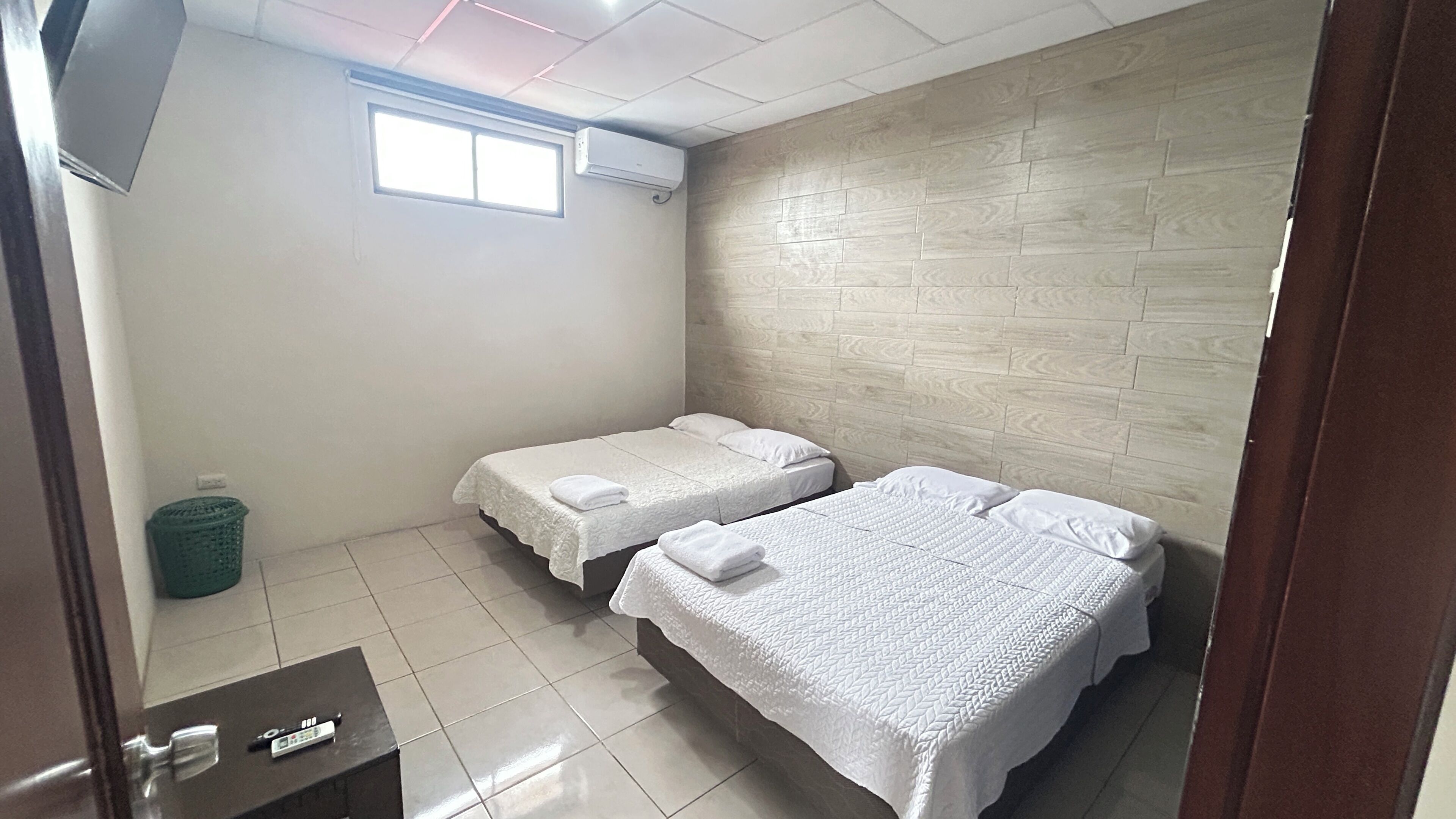 A hotel room with two beds, a window, a trash bin, and a control panel.