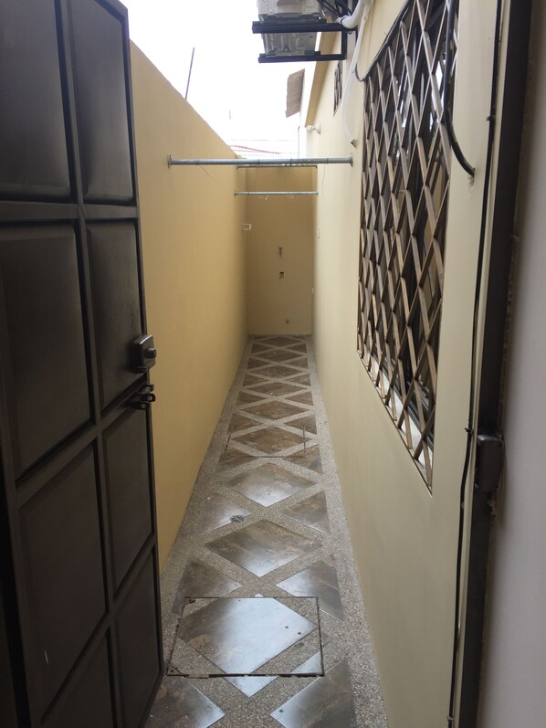 A narrow hallway with a tiled floor, a metal door on the left, and a barred window on the right. - NCG Suite 1 (Guayaquil)