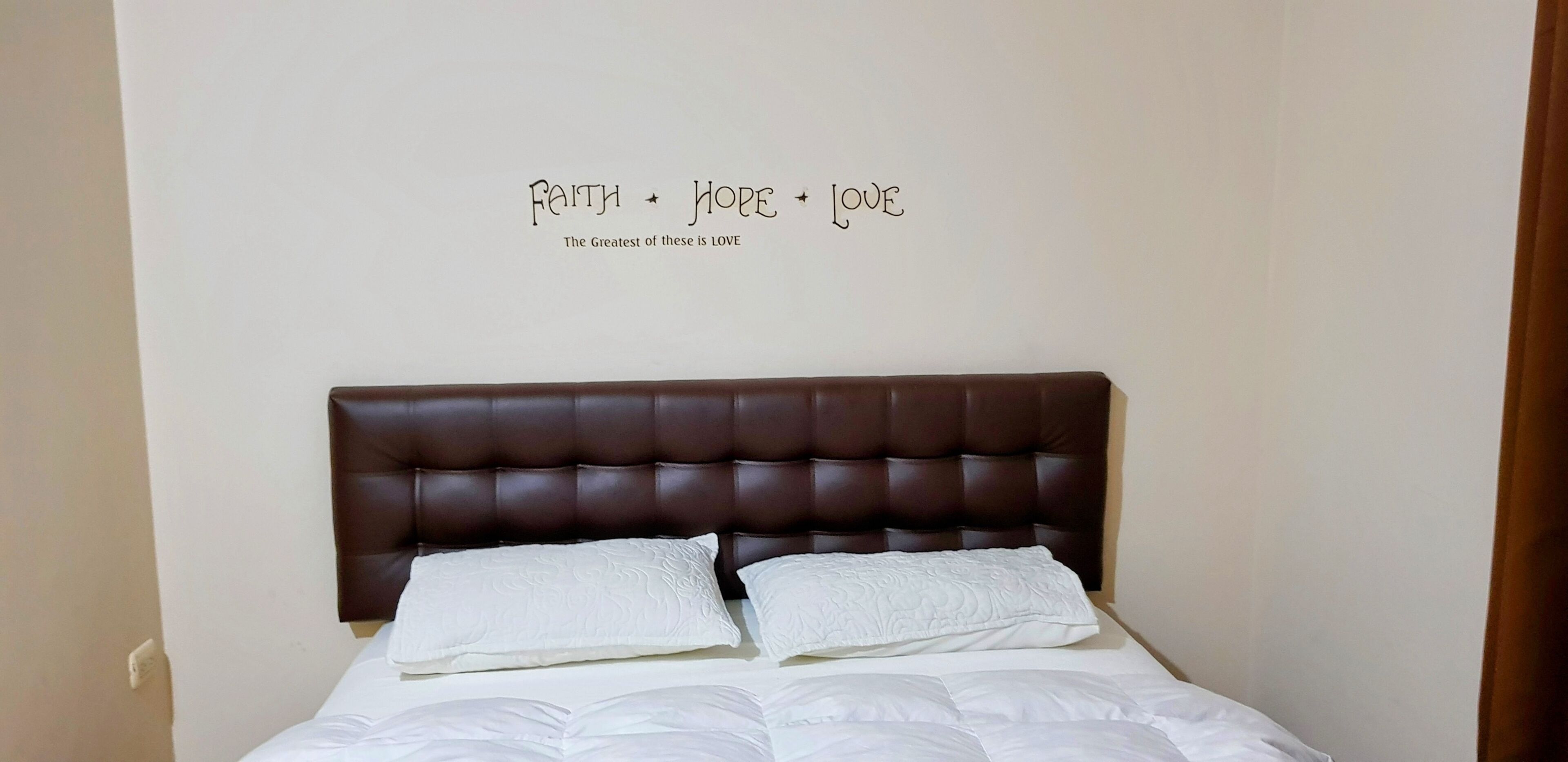 A neatly made bed with a brown headboard, white bedding, and a wall with inspirational words.