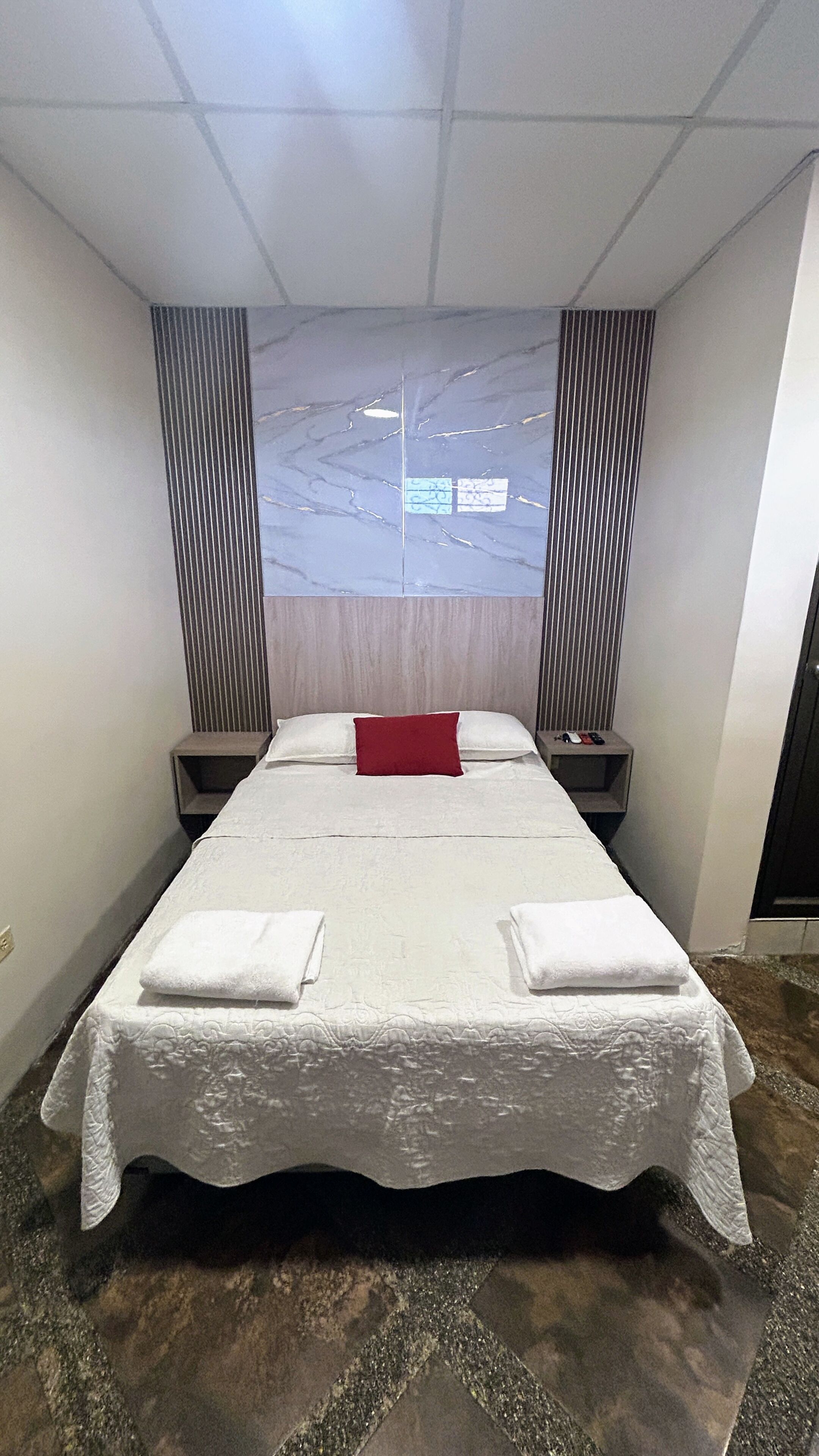 A hotel room with a large bed, two bedside tables, a red pillow, and a view of the sky through a window.