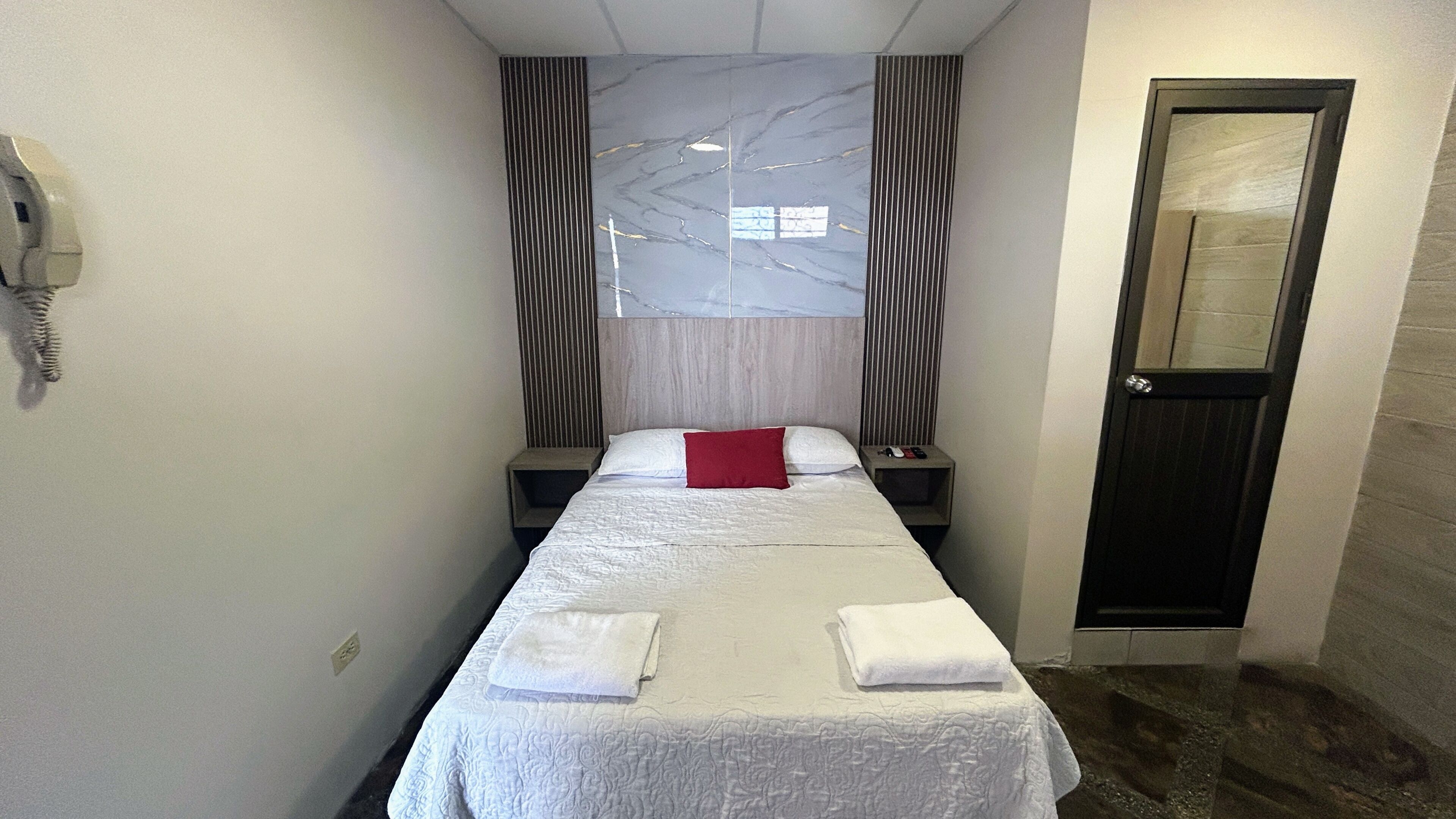 A hotel room with a bed, a bedside table, a red pillow, a white towel, a telephone, and a door.