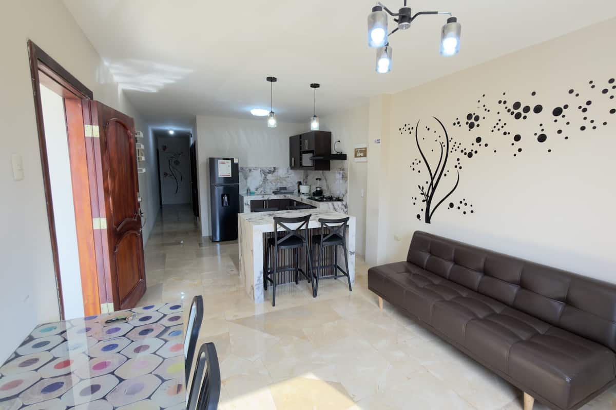 A modern living room with a brown sofa, a dining table with colorful chairs, a kitchen with dark cabinets, and a decorative wall mural.