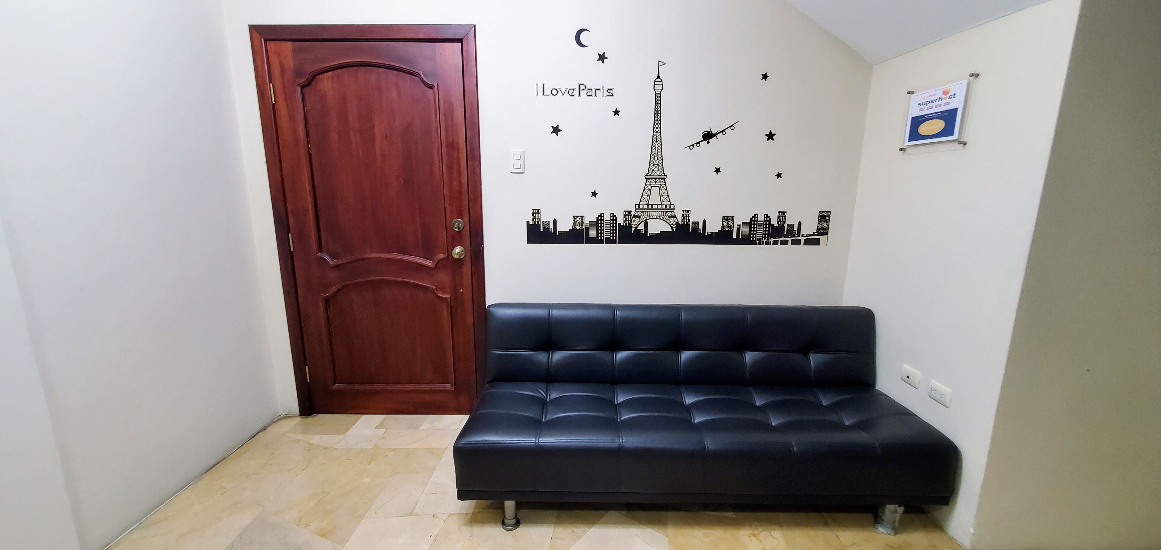 Una habitació amb una porta de fusta, un sofà negre, una samarra mural de monuments de París i un cartell al mur.