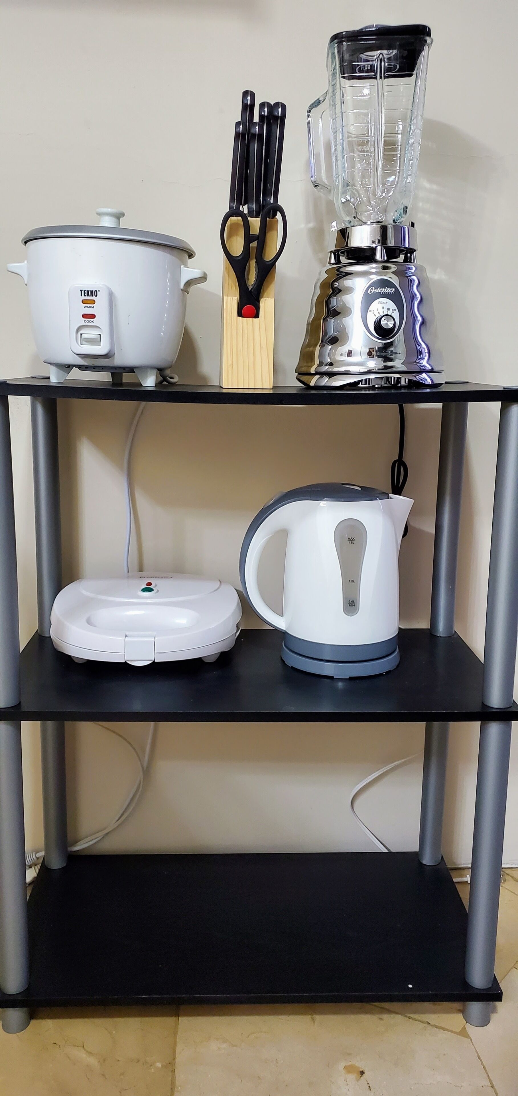 A kitchen shelf with a slow cooker, a blender, a kettle, and a pair of scissors.