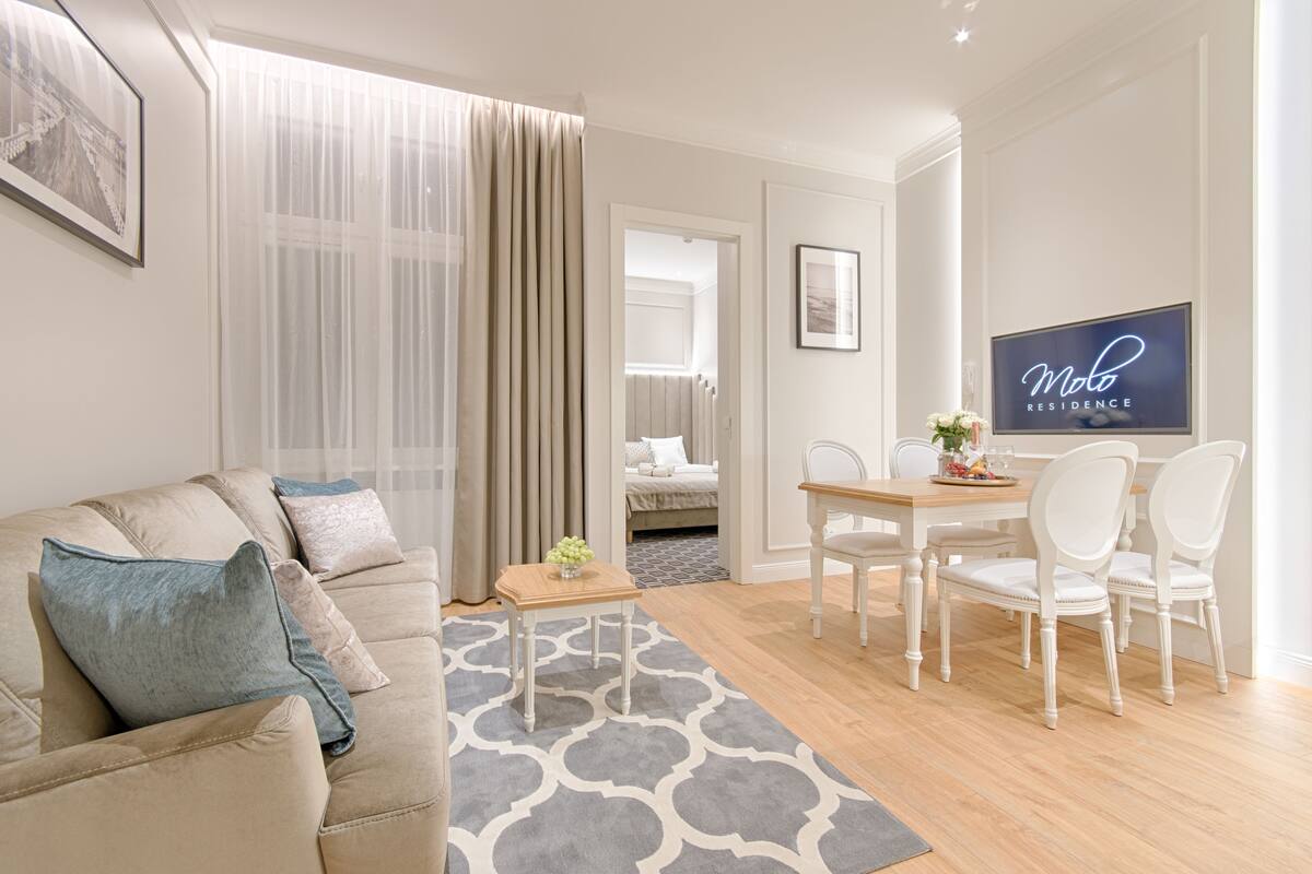 Un salon moderne avec un canapé beige, une table à manger avec des chaises blanches, un tapis à motifs gris et un téléviseur affichant l’image de « Molo Residence ».