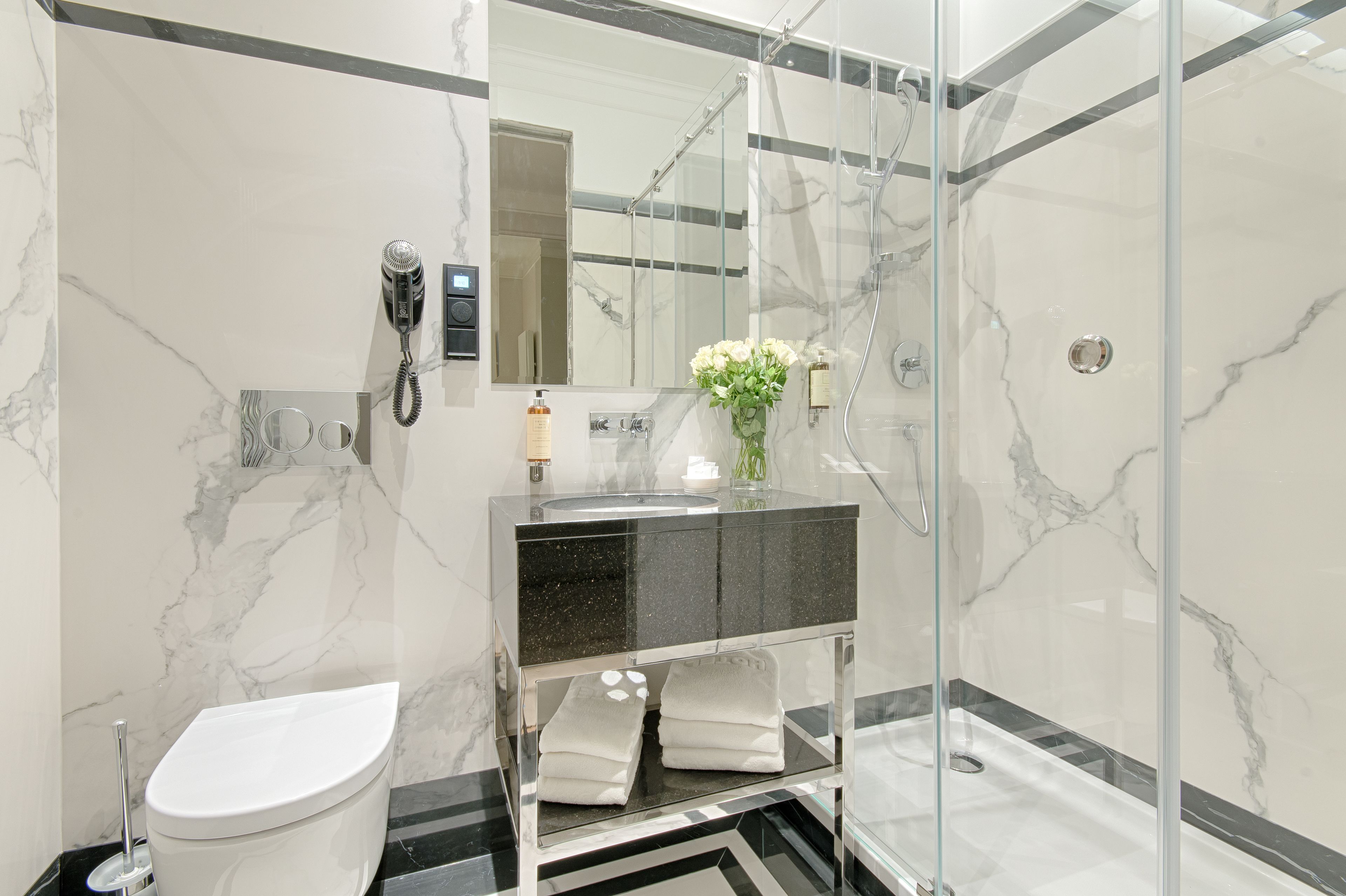 A modern bathroom with a glass-enclosed shower, a marble-tiled wall, a black and white vanity, and a toilet.