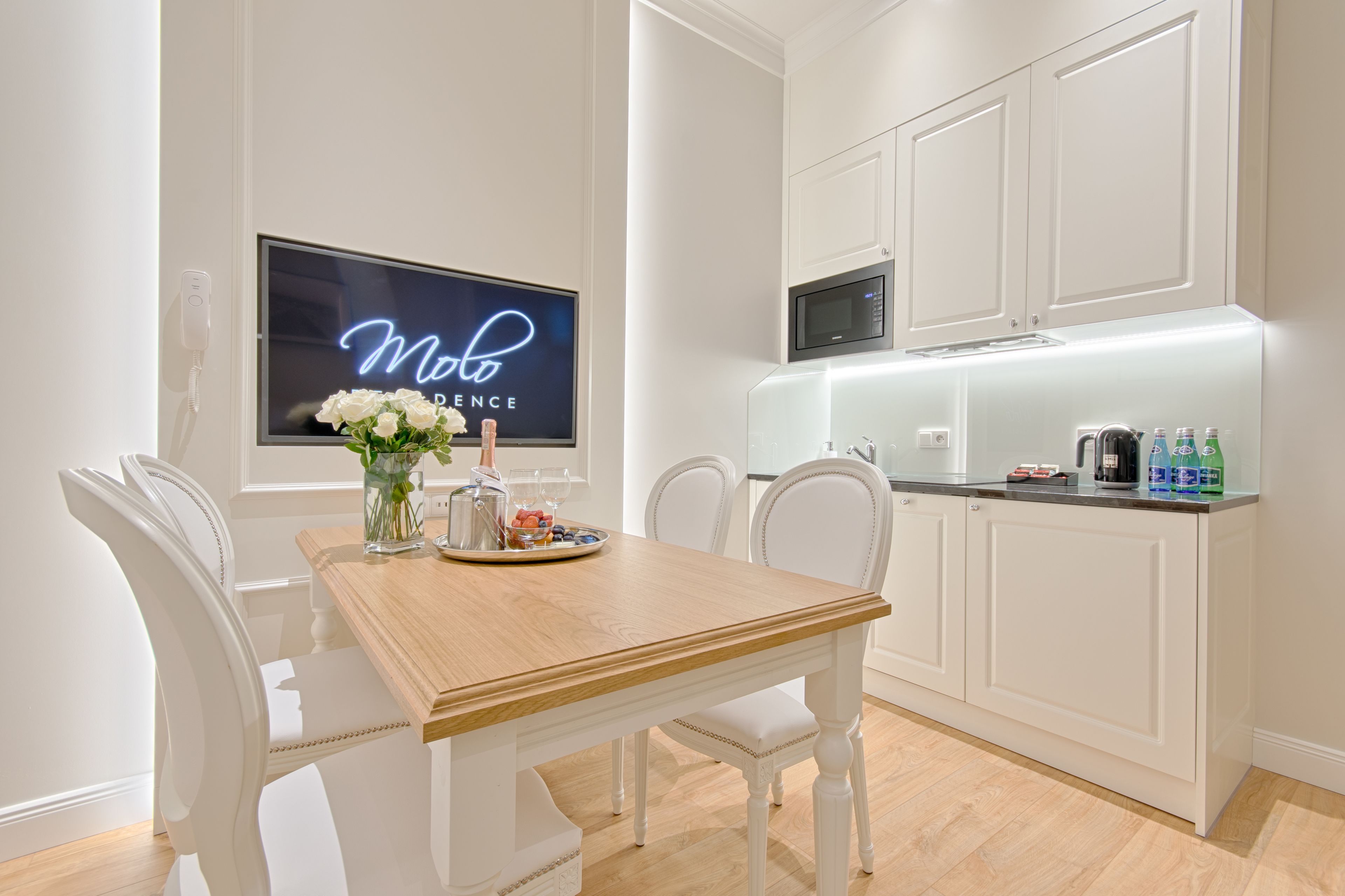 A modern kitchen with white cabinetry, a wooden dining table, and a neon sign on the wall.
