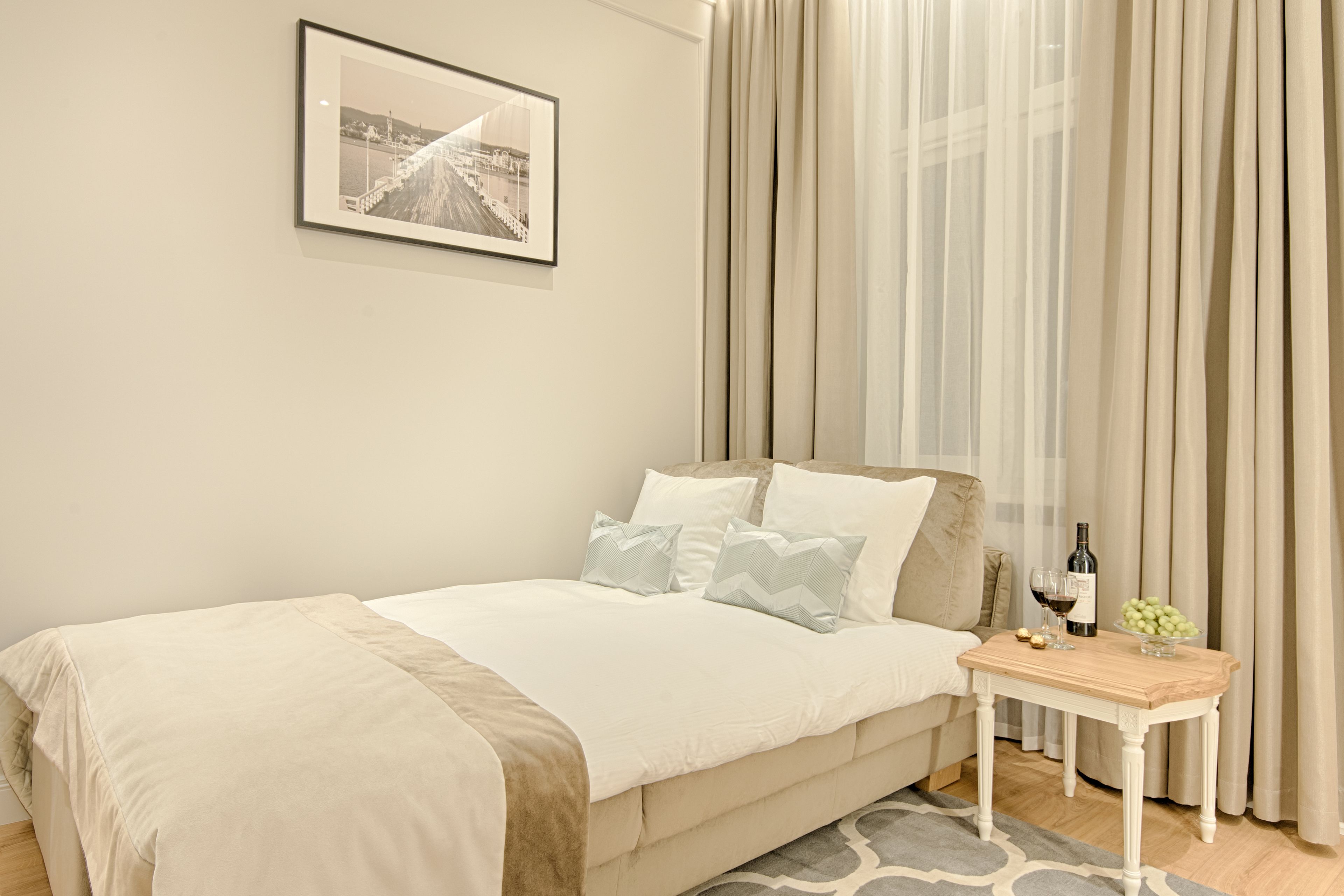 A neatly made bed with a beige cover, a small bedside table with a bottle and glasses, and a framed picture on the wall.