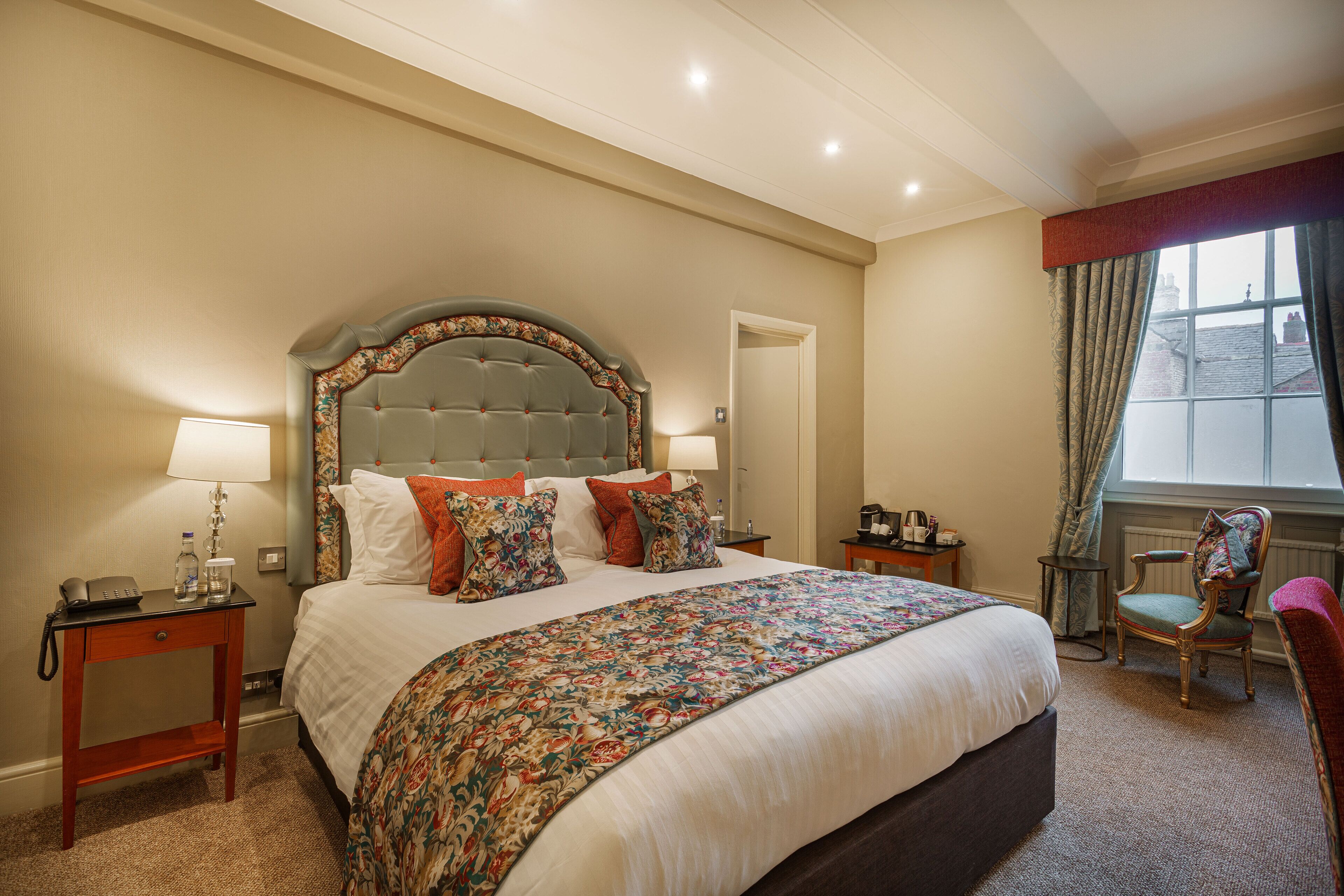 Habitación de hotel con una cama grande, una mesita de noche con lámpara, una silla y vistas al exterior a través de una ventana.