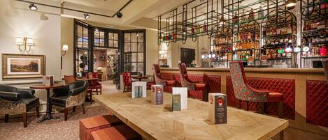 Een bar met een houten tafel, rode fauteuils en diverse flessen op planken.
