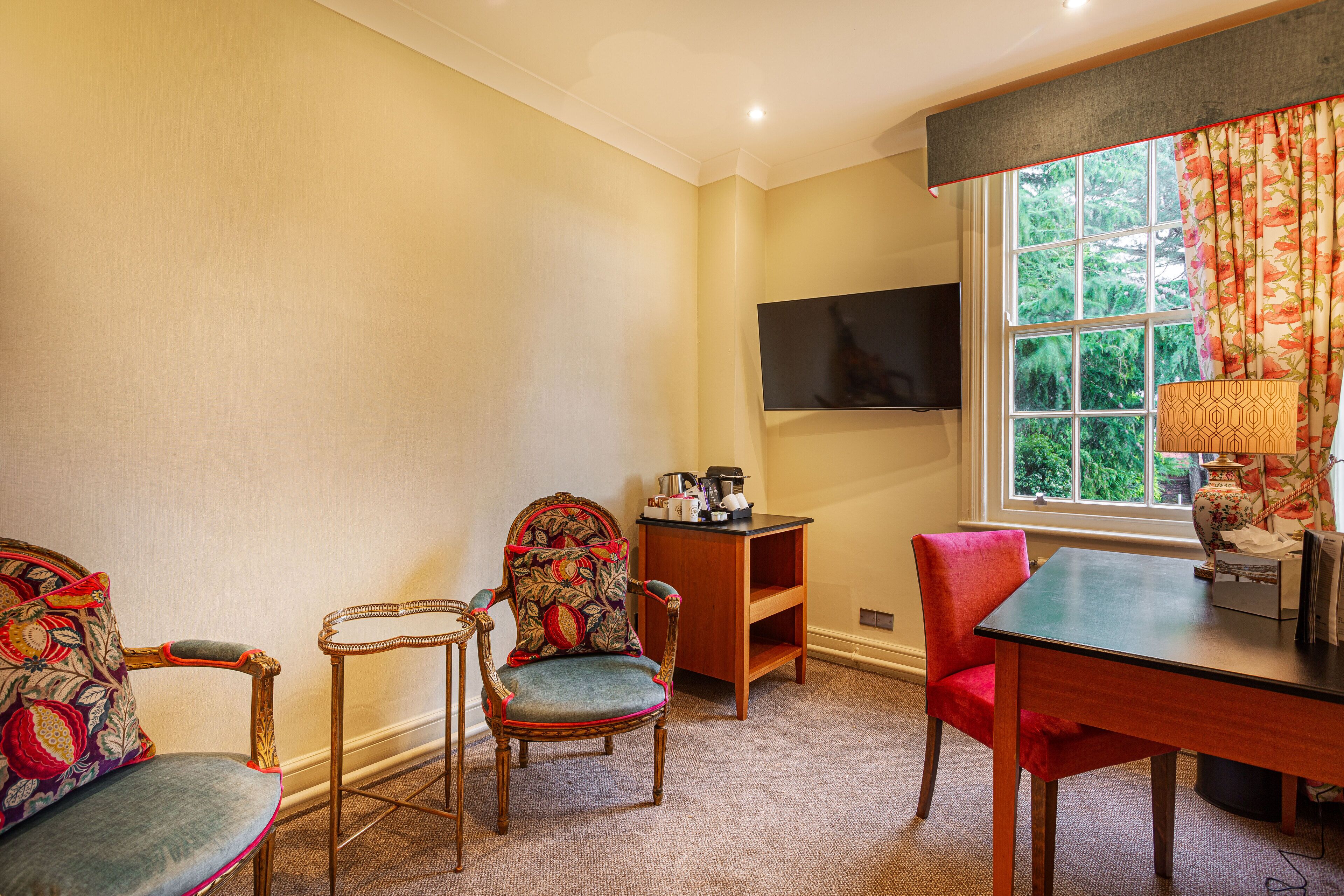 A room with a TV mounted on the wall, a wooden table, two chairs, and a window with curtains.
