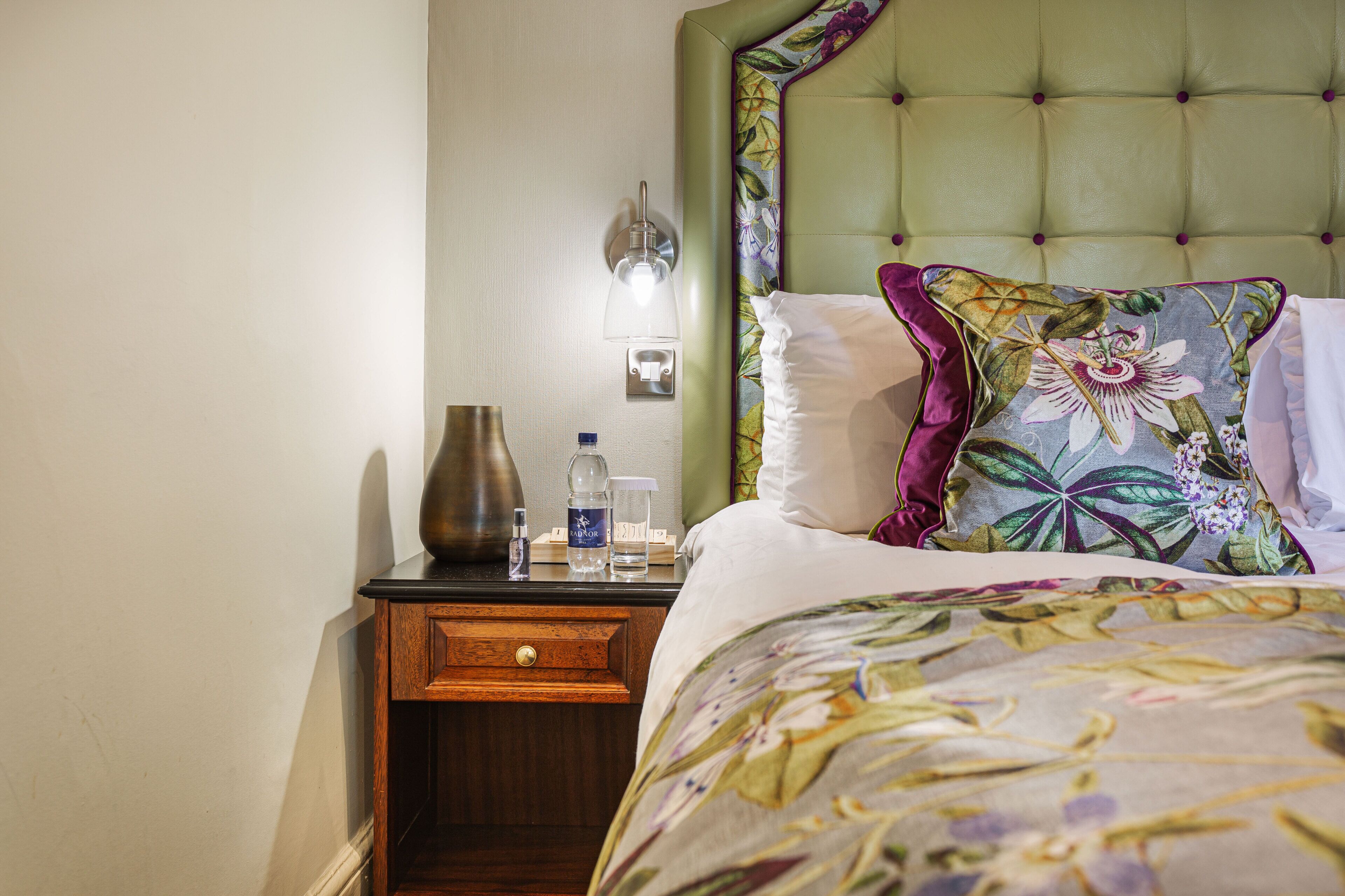 A hotel room with a bed, a nightstand, a lamp, a vase, and a bottle.