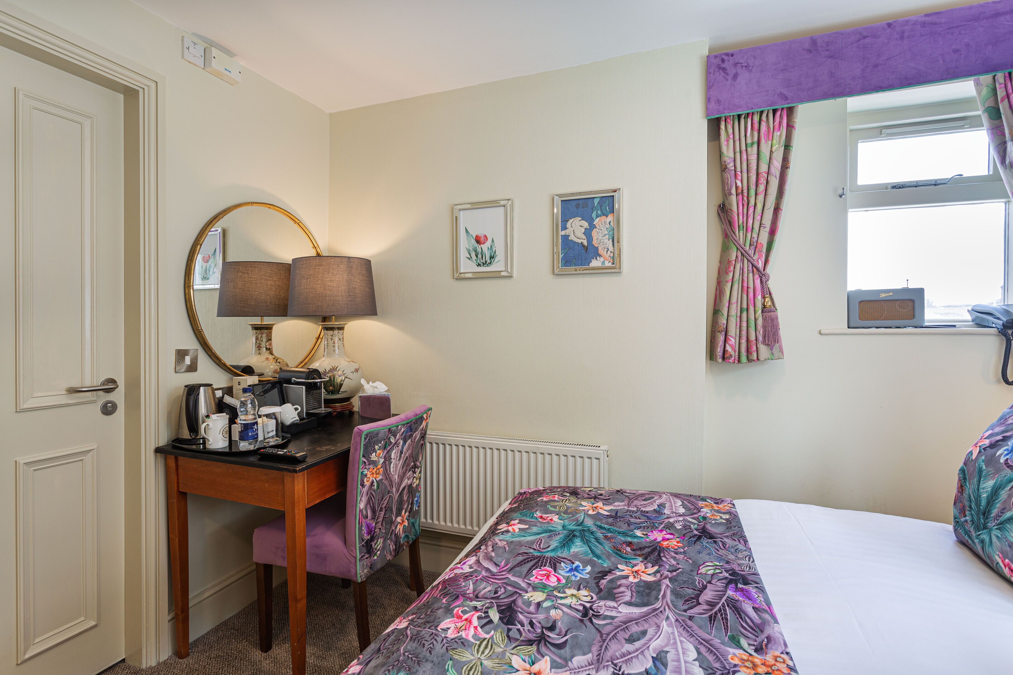 Habitación de hotel con una cama, un escritorio con lámpara, una silla, un radiador, un ventanal con cortinas y dos cuadros en la pared.
