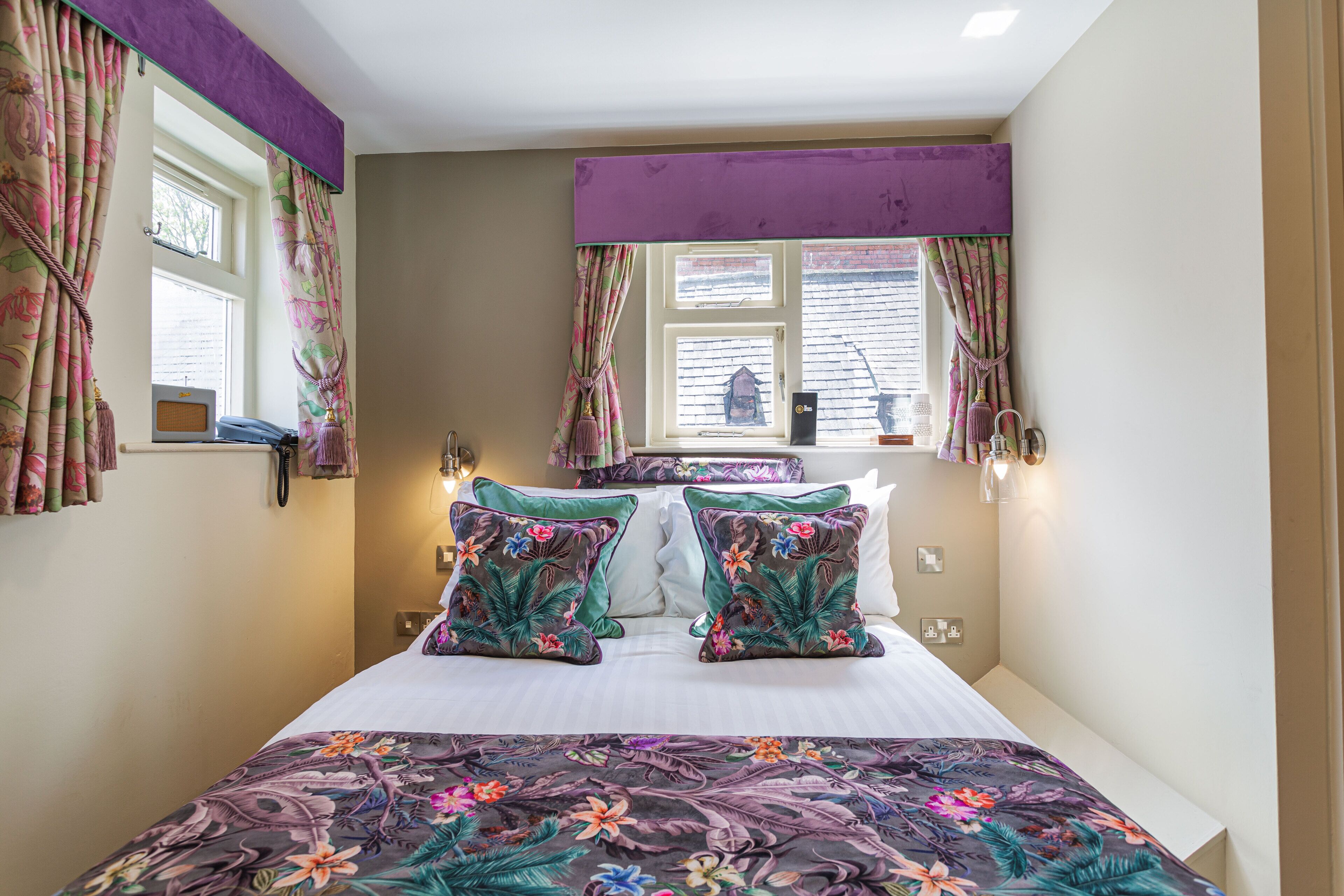 Une chambre d’hôtel avec un lit, des rideaux à motifs floraux, une fenêtre donnant sur un beau paysage et une lampe.