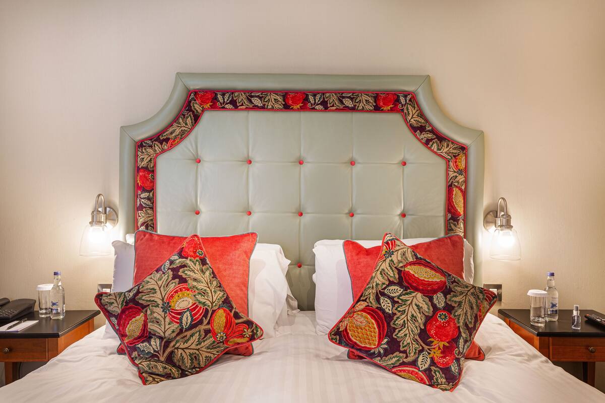 Ein Hotelzimmer mit einer gepolsterten Kopfteil, Kissen mit Blumenmuster, einem Nachttisch mit Lampe und einem Beistelltisch mit Telefon und Flasche.
