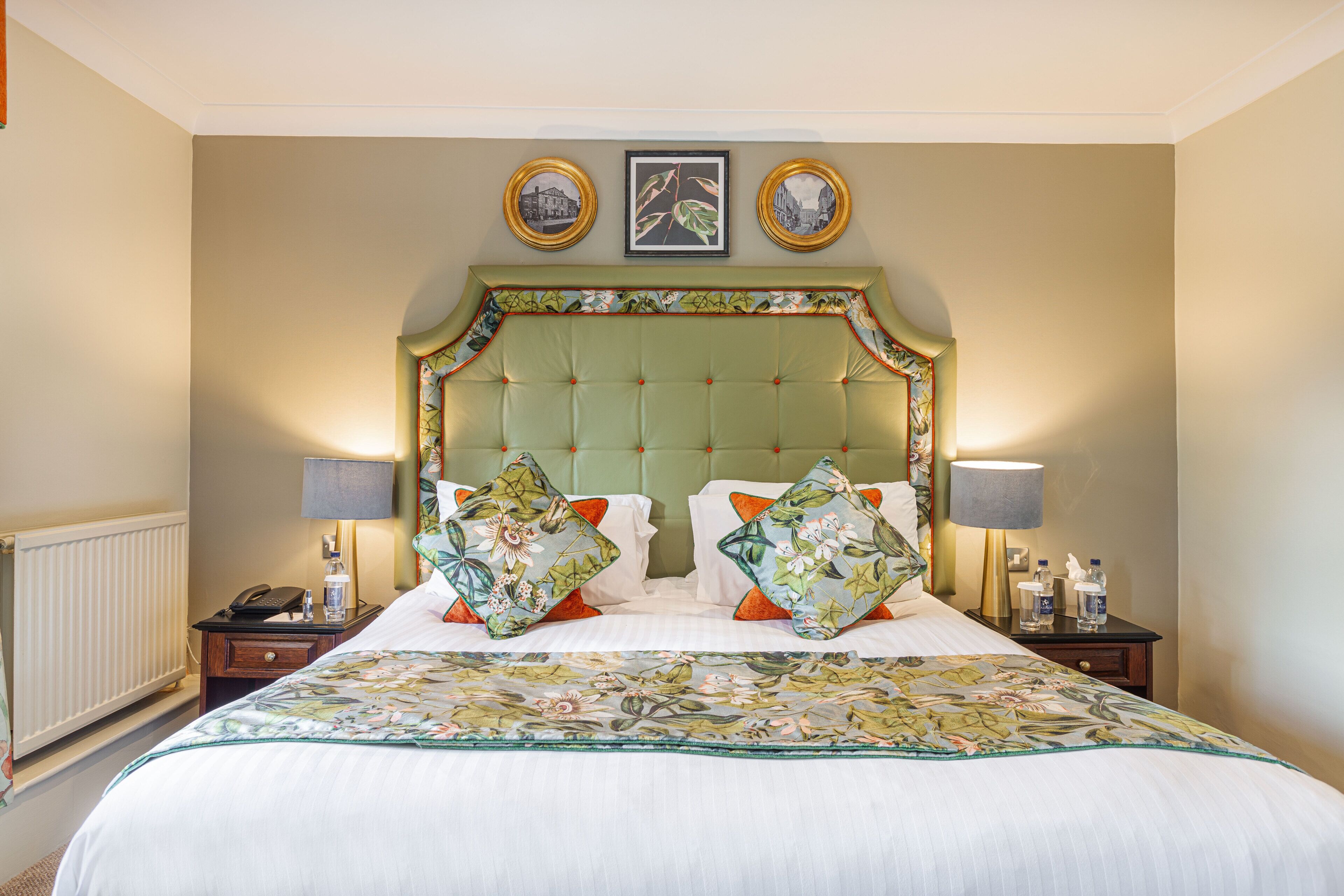 A hotel room with a large bed, two bedside tables, a headboard with a floral pattern, and two framed pictures above the bed.