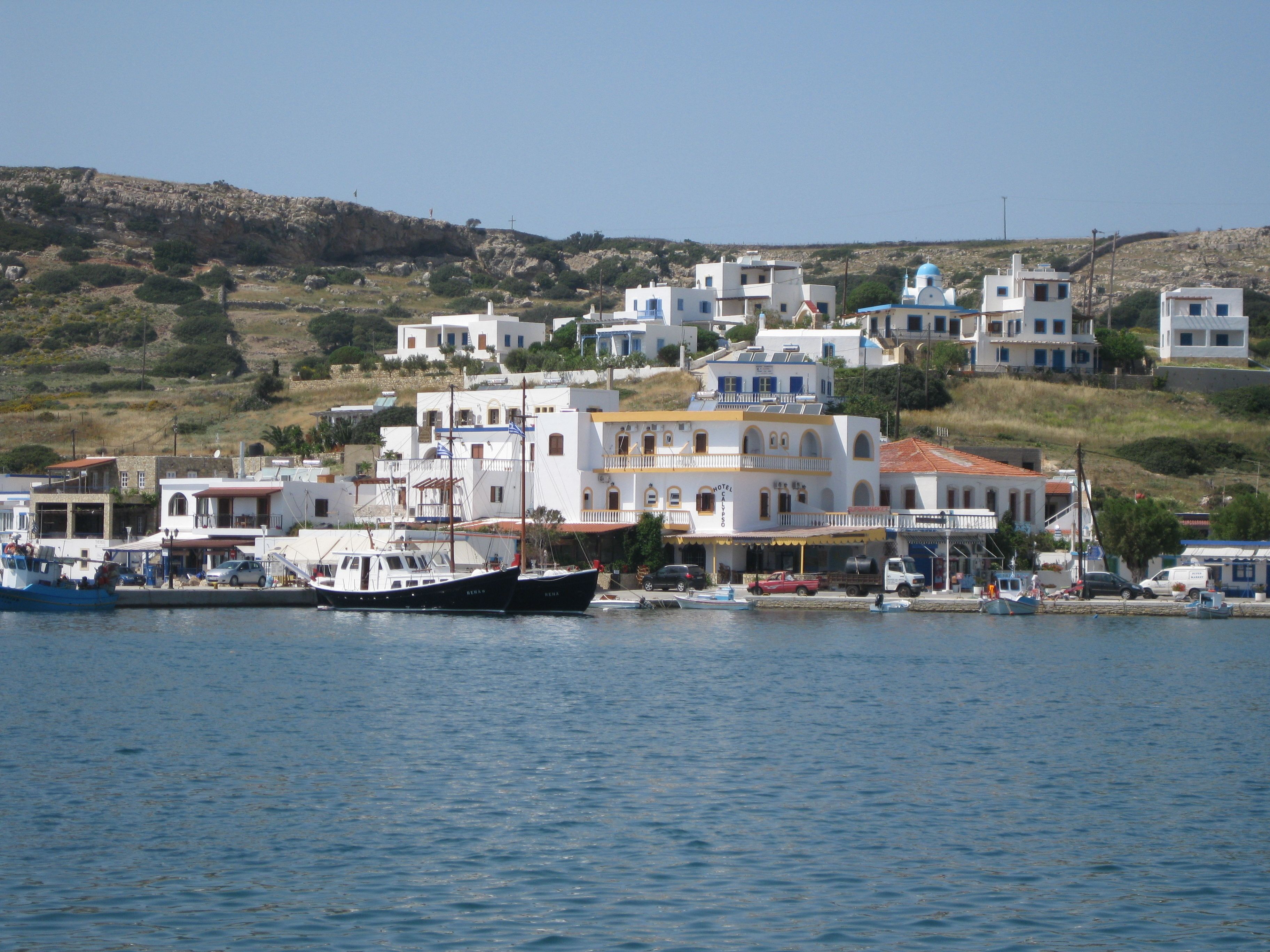 Un pueblo costero con edificios blancos, un muelle con barcos y una colina al fondo.