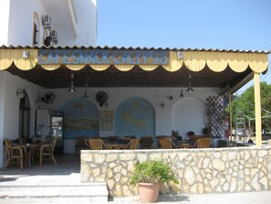 Una cafetería con mesas al aire libre, paredes de piedra y un letrero que dice "CAFE SNACK CALYPSO".