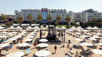 Una platja amb gandules i ombrelles, un pavelló central i un edifici d'hotel al fons.