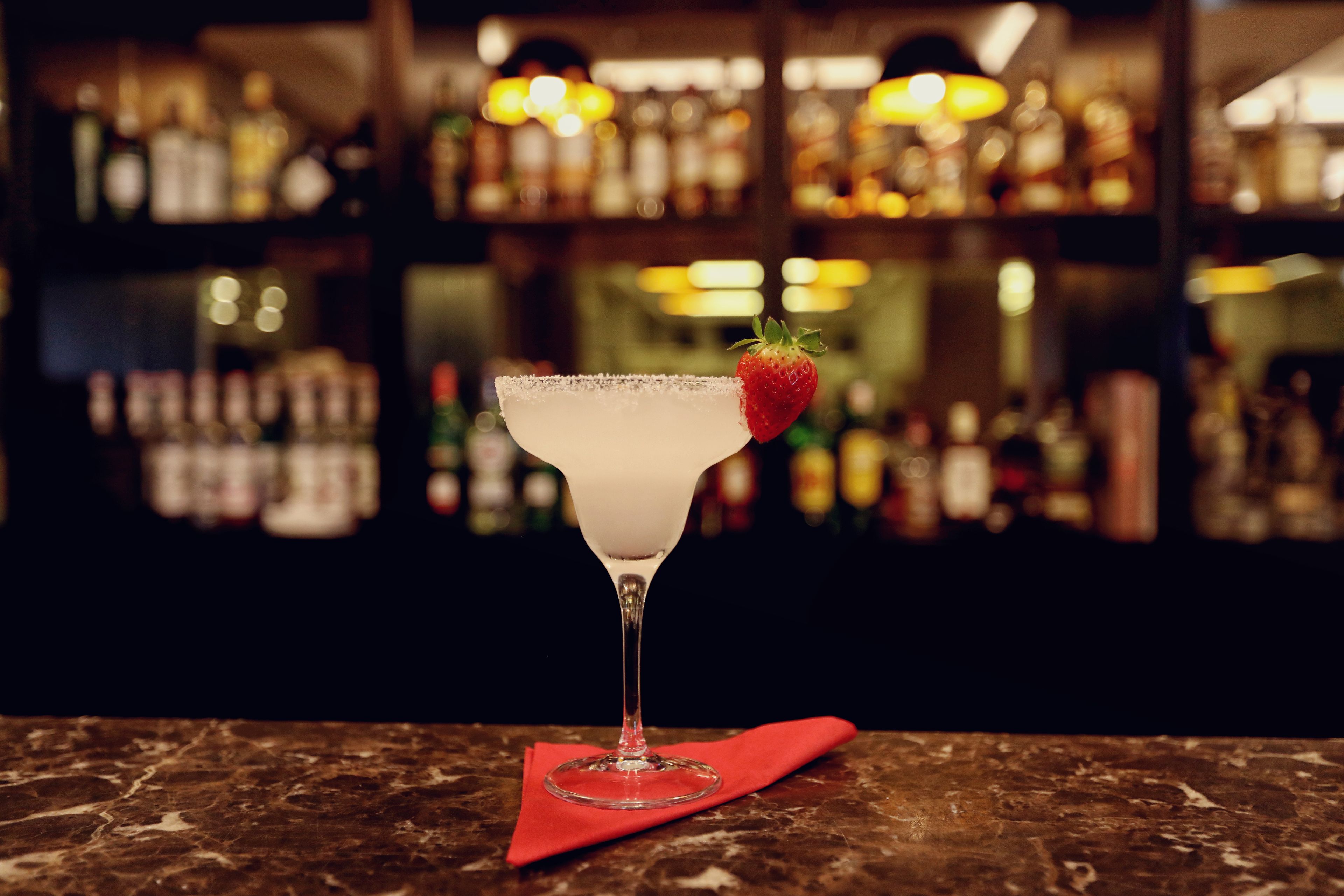 A martini glass with a strawberry garnish on a marble surface, with a blurred bar in the background.