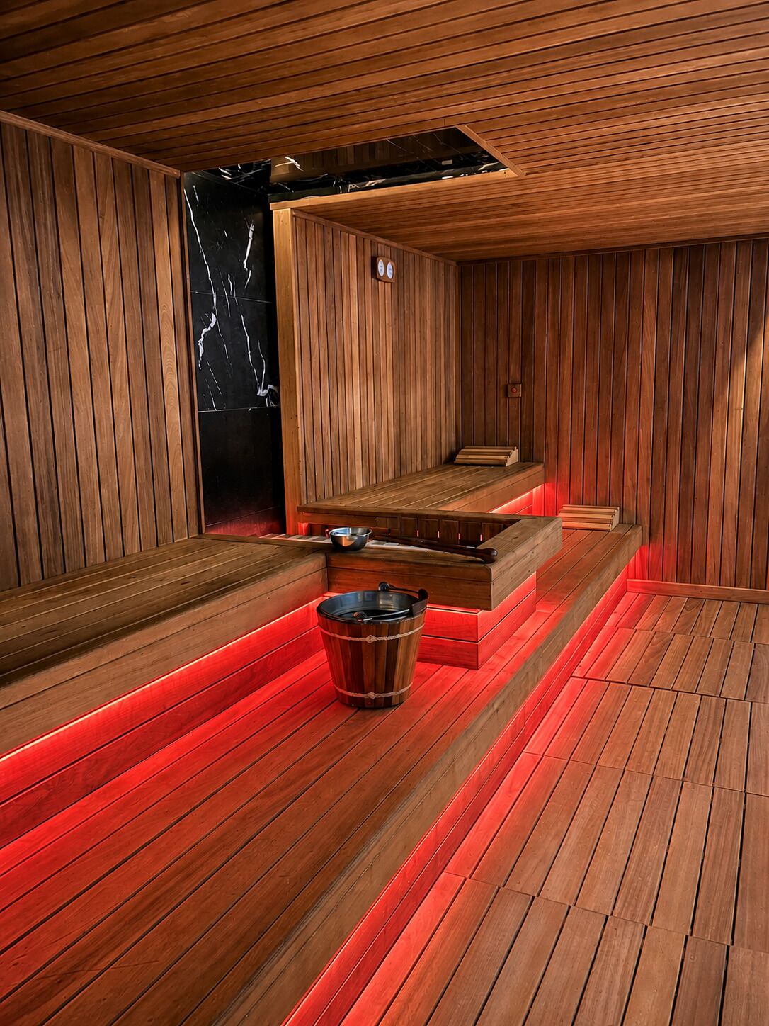 A sauna room with wooden walls and benches, a red-lit floor, and a metal bucket.