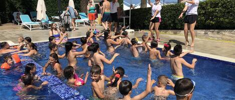 Piscina com crianças e adultos aproveitando a água, cercada por espreguiçadeiras e guarda-sóis.