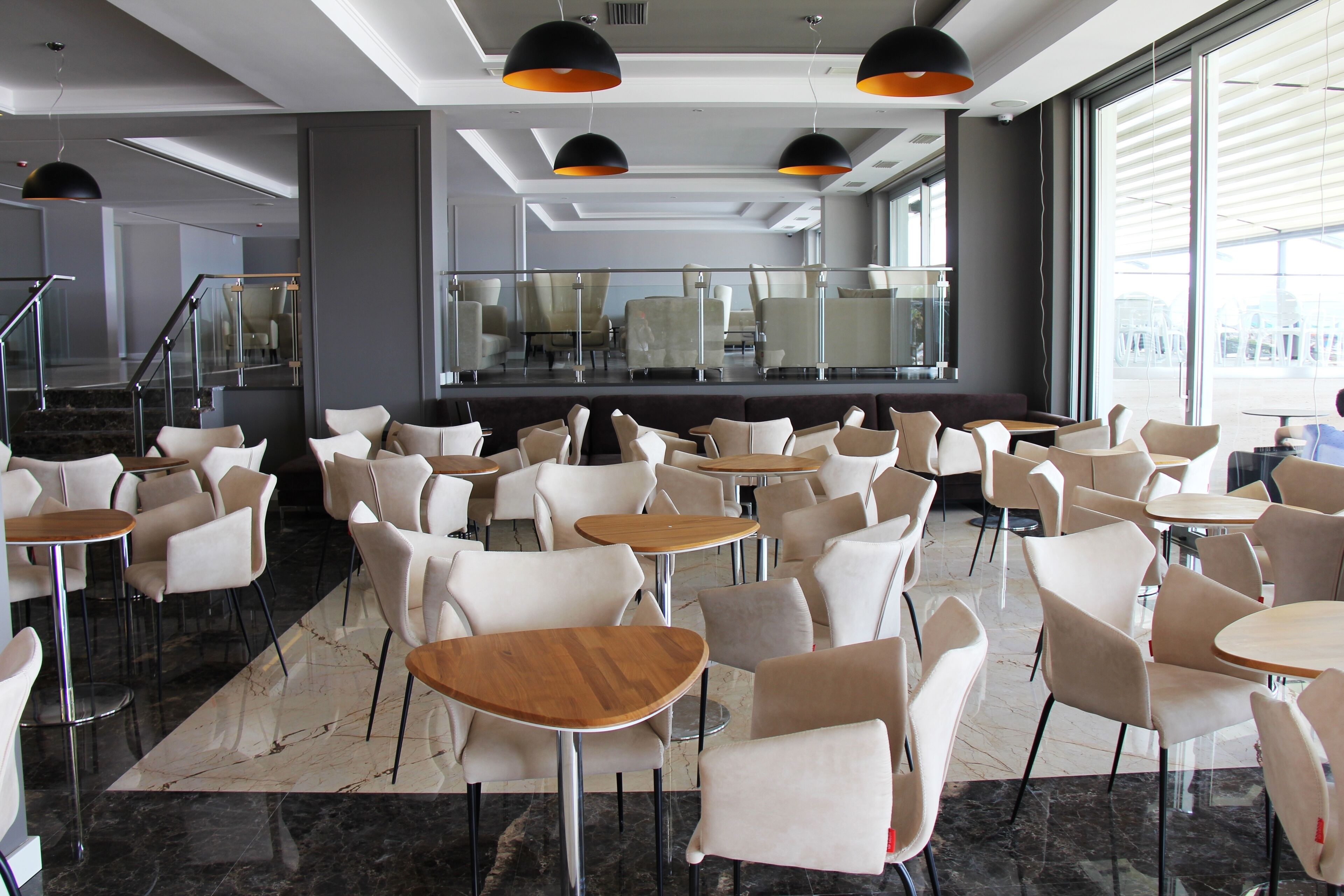 A modern cafe with multiple seating areas, including round tables and cushioned chairs, arranged in a spacious layout with large windows offering views of the outside.