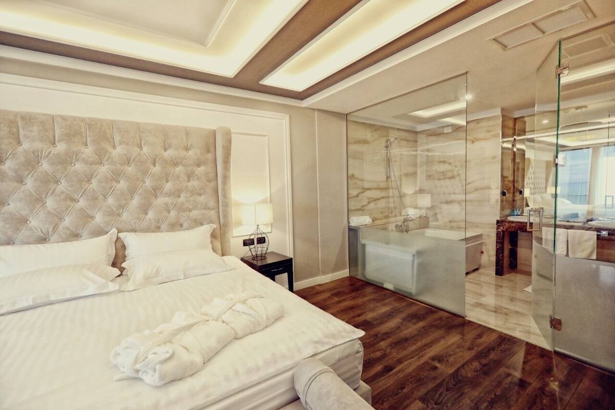 A modern hotel room with a large bed, a glass-enclosed bathroom, and a wooden floor.
