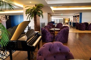 A lobby with a grand piano, plush purple seating, a palm tree, and a modern interior design. - Premium Beach Hotel (Golem)