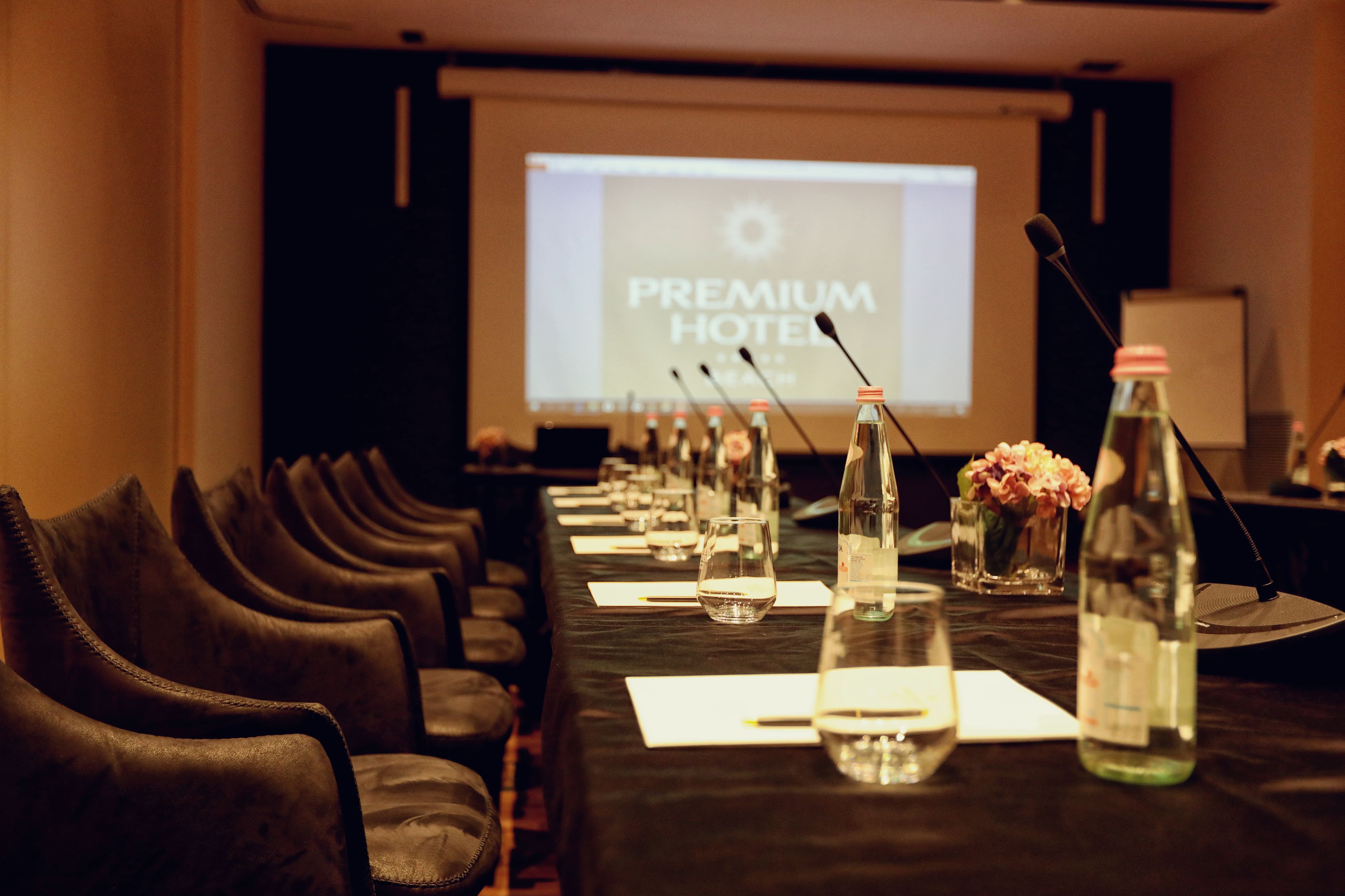 A conference room with a long table, chairs, microphones, water bottles, and a screen displaying "PREMIUM HOTEL".