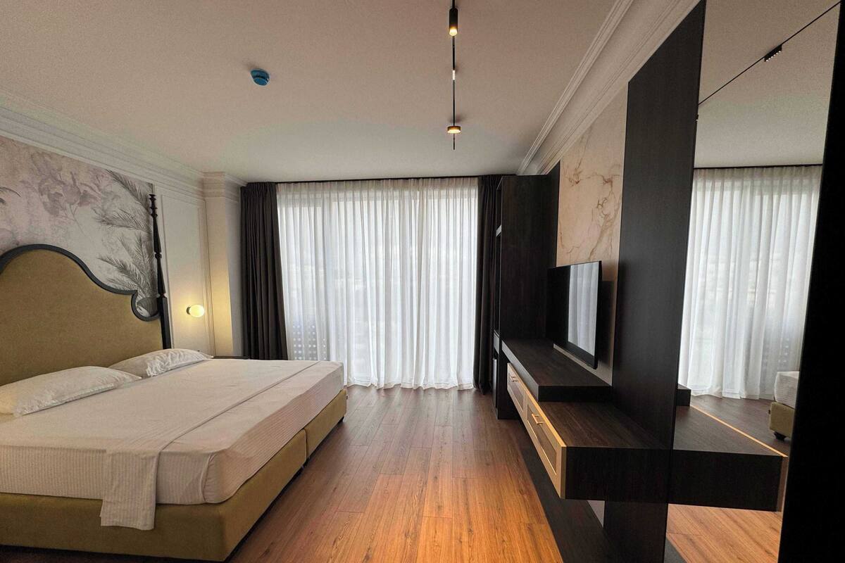 A hotel room with a bed, a TV, a desk, and a large window with curtains.