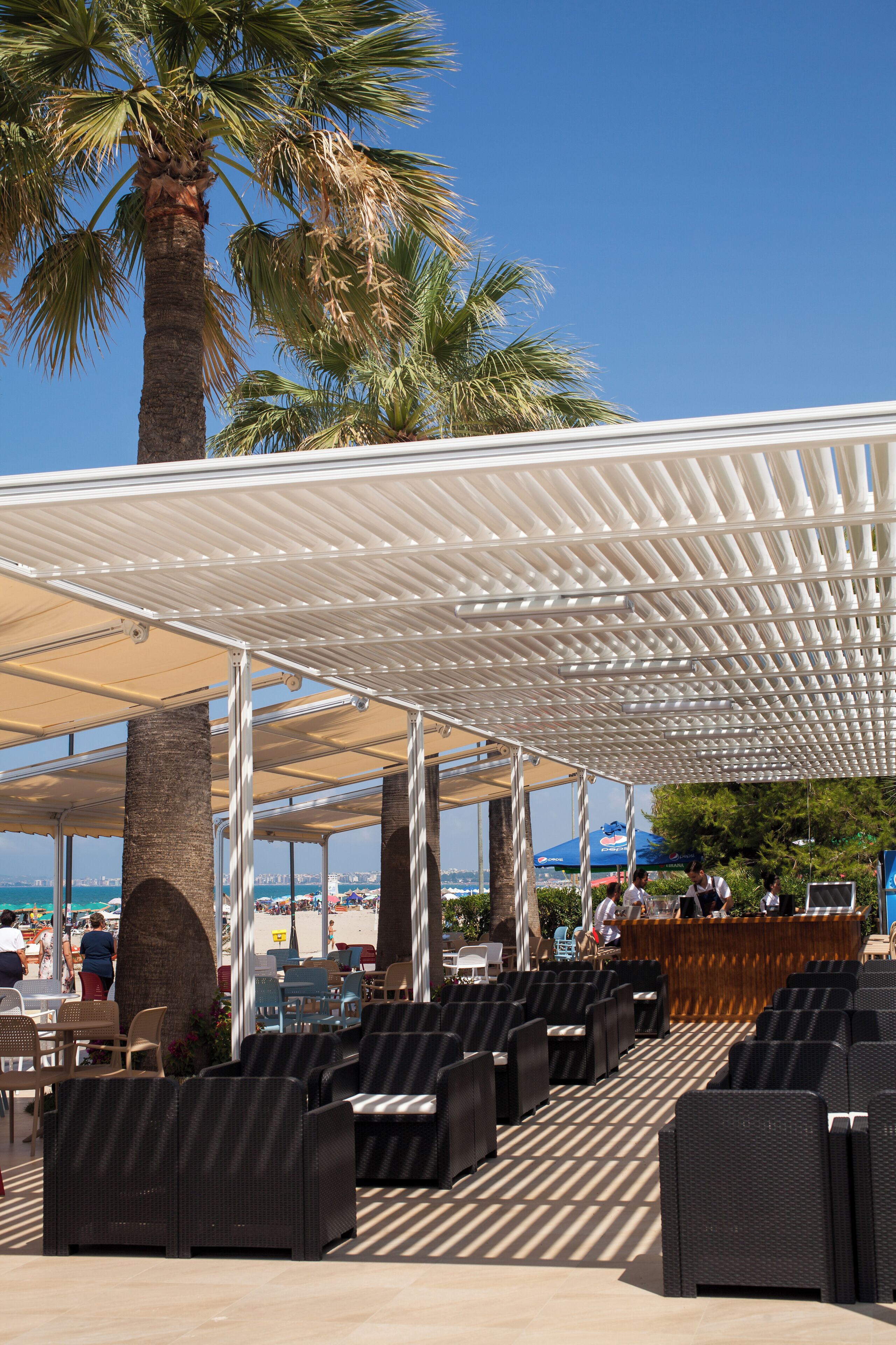 An outdoor seating area with wicker chairs and a covered section, palm trees, and a view of the beach.